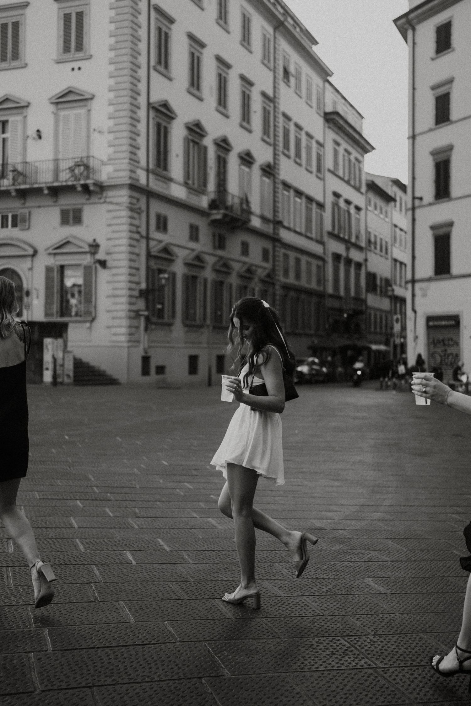 Young woman in white dress walking through a classic European city square, holding a cup in black and white photo.