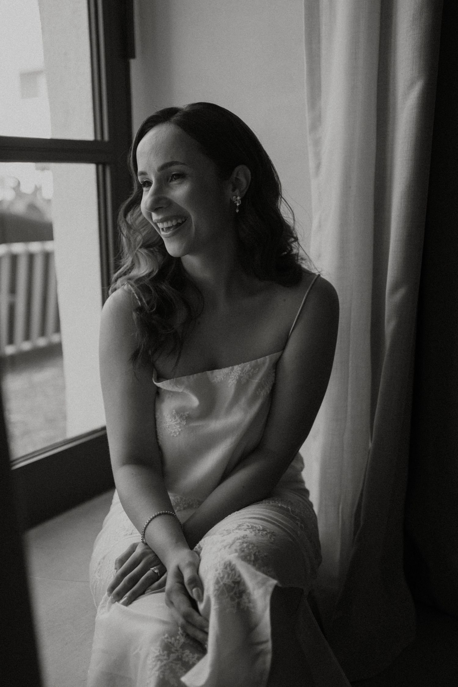 Joyful bride in white slip dress laughing by a window in black and white portrait.