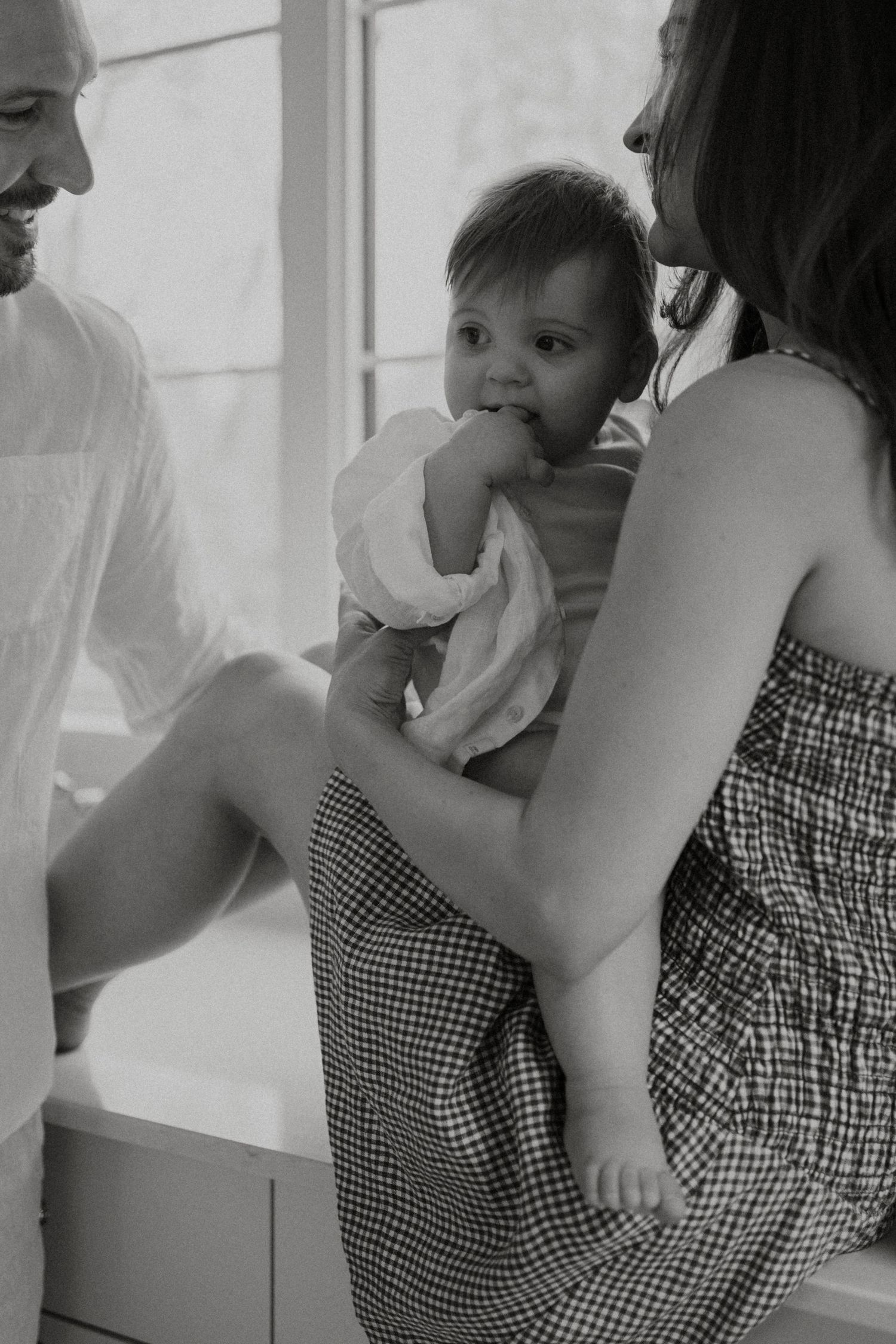 Black and white photo of a mother holding her baby wrapped in a towel, sitting beside a smiling man near a window.