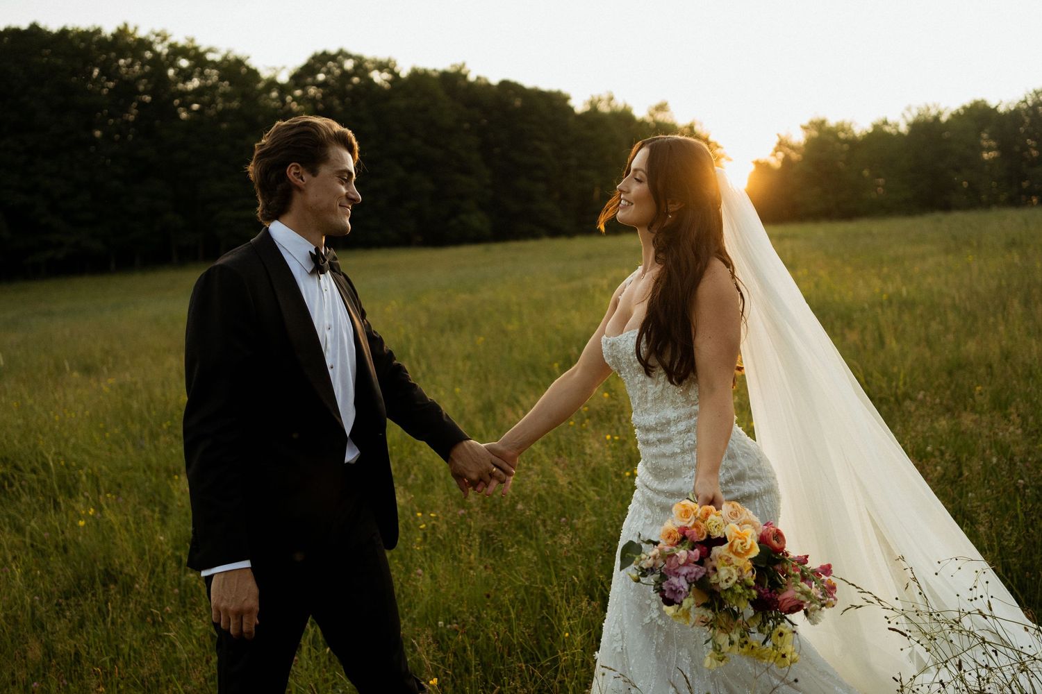 Bride and groom holding hands in golden sunset field, bride wearing white gown and veil holding colorful bouquet.