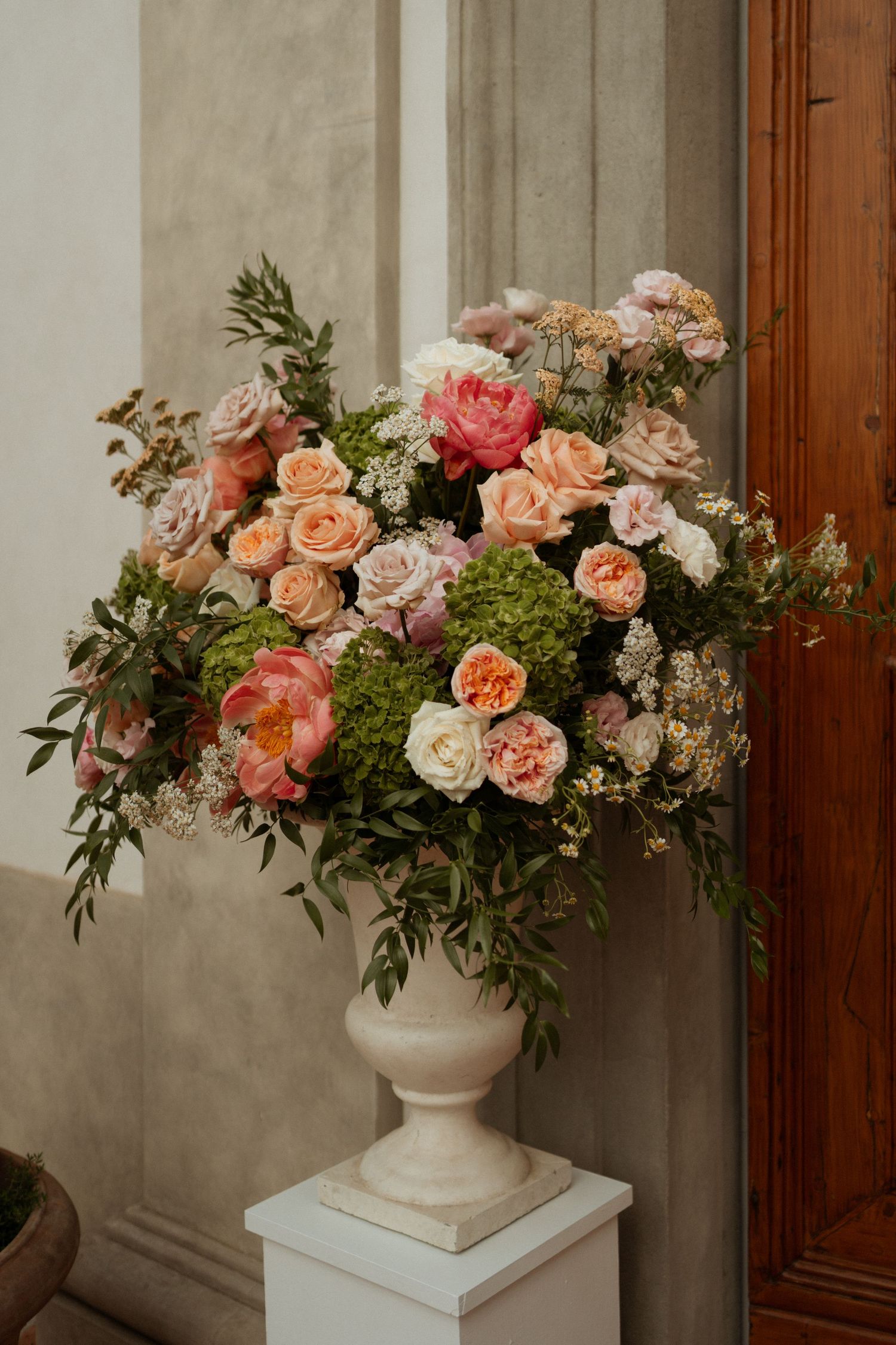 Elegant floral arrangement of pink peonies, peach roses, and greenery in white urn vase.
