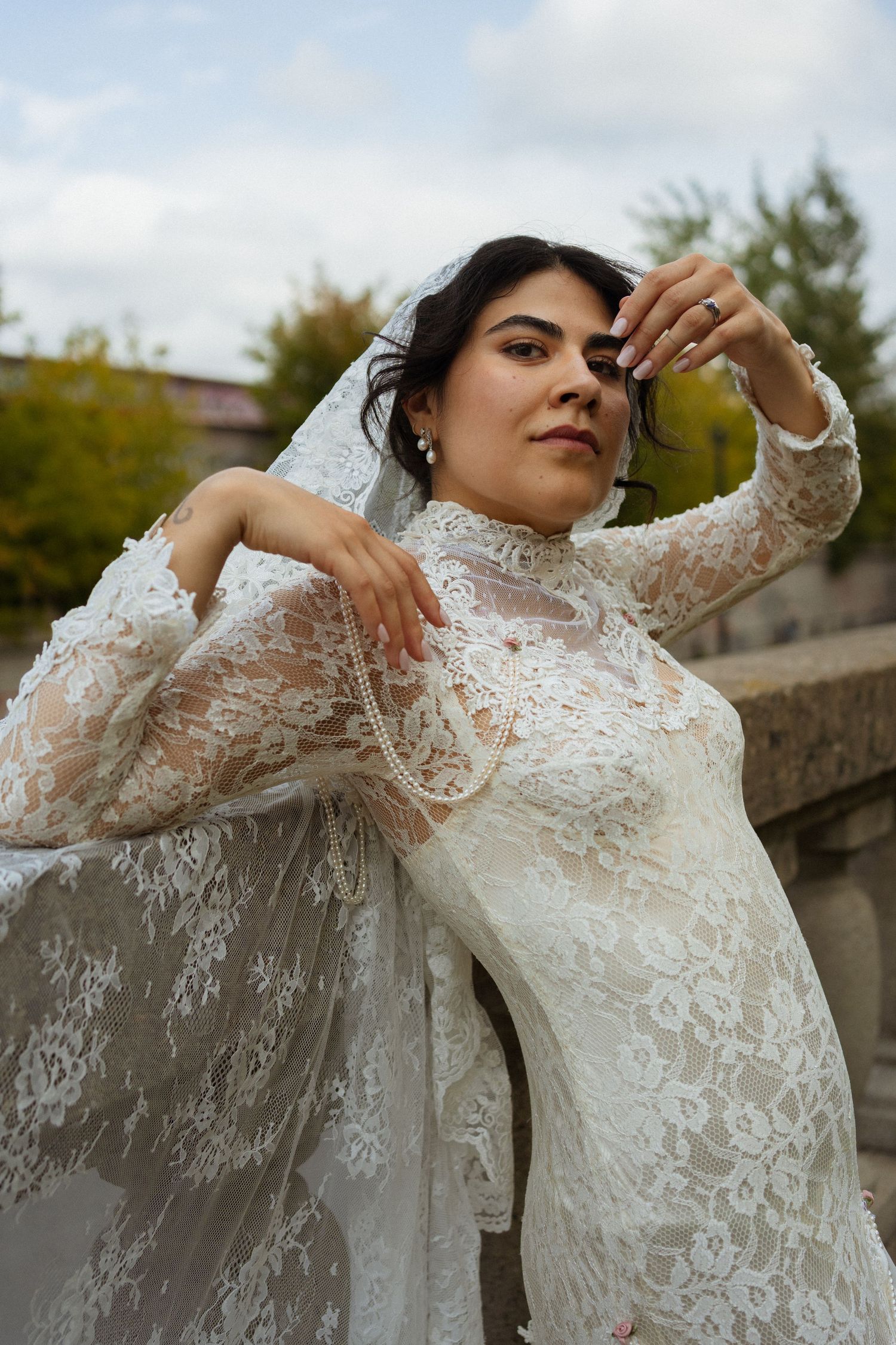 A bride in a white lace gown and veil poses gracefully outdoors, showcasing elegant bridal fashion.
