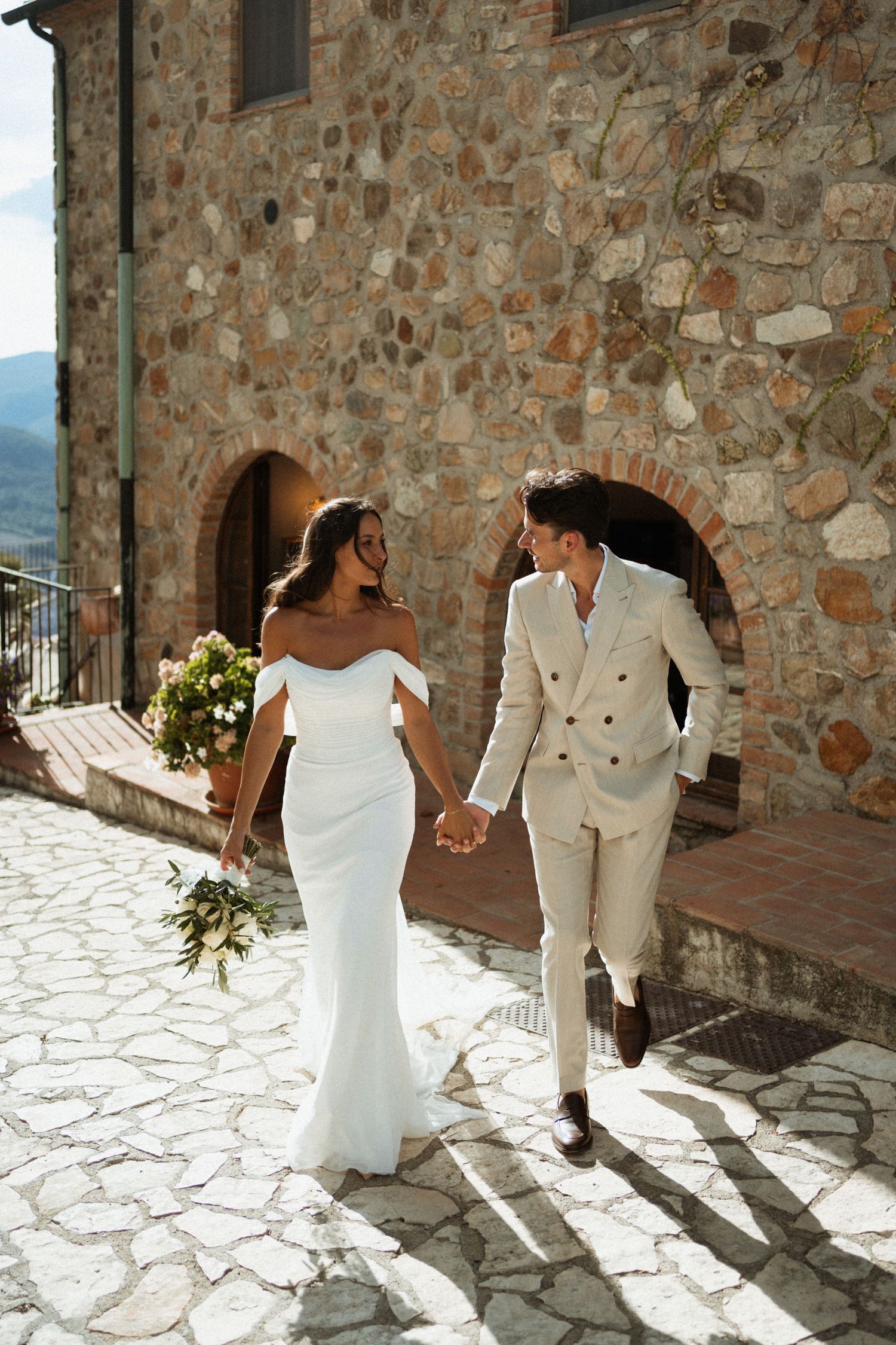 Happy bride and groom holding hands walking on cobblestone path beside a rustic stone building.