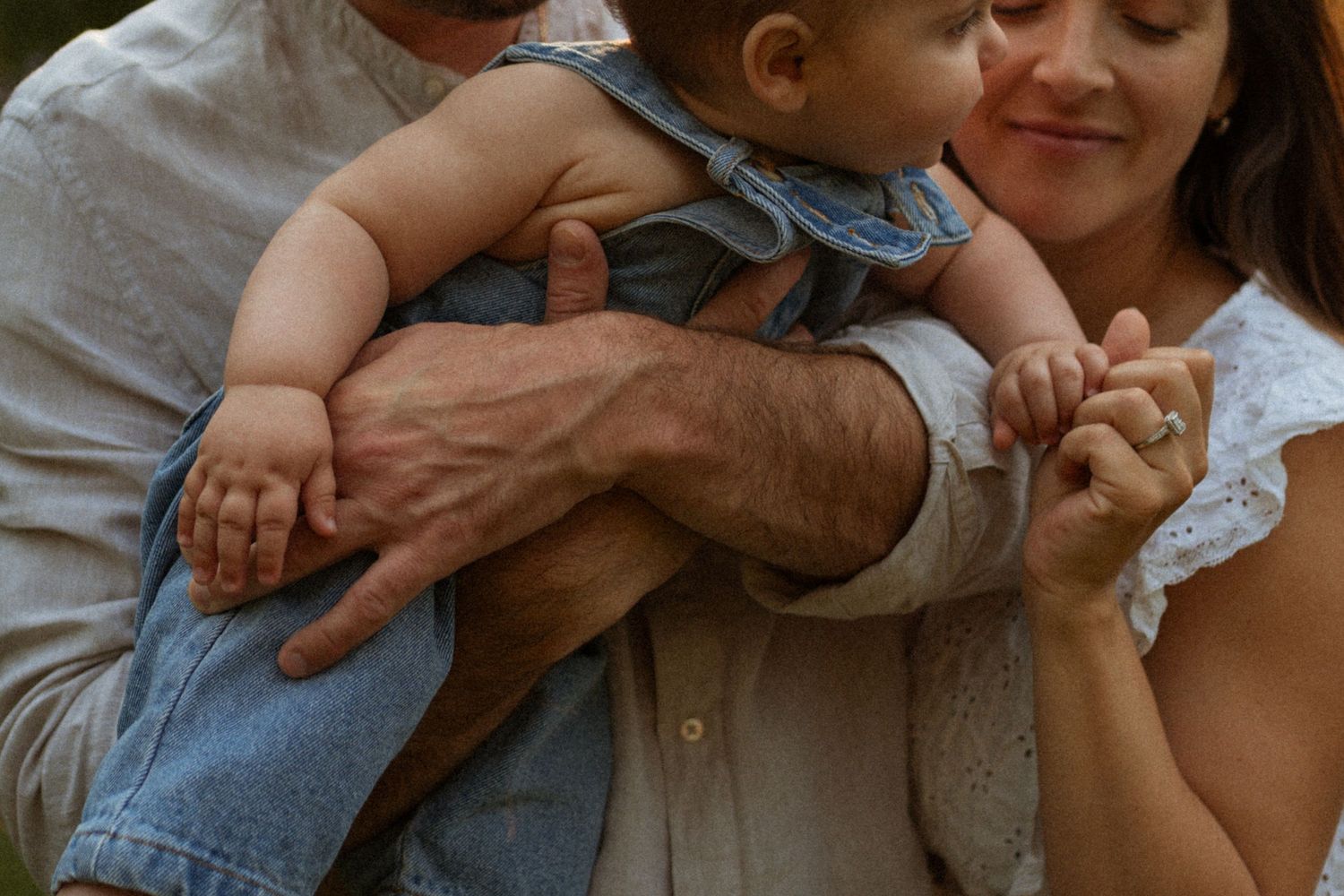 Parents tenderly hold their baby in denim outfit outdoors, mother smiling softly in white lace dress.