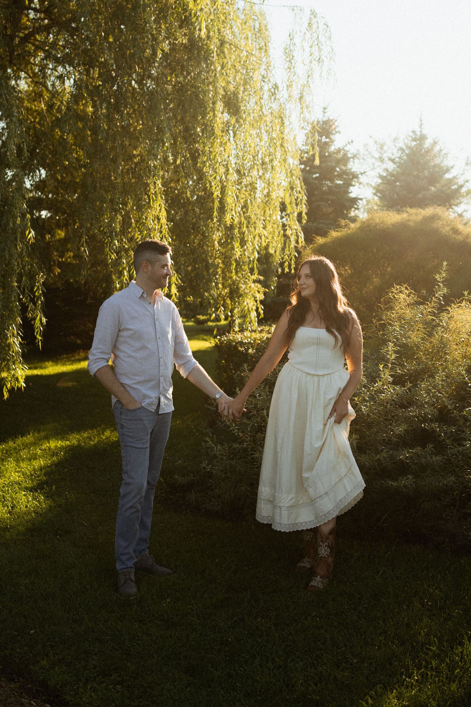Couple holding hands in golden hour sunlight beside a weeping willow tree, woman in white dress, man in white shirt.