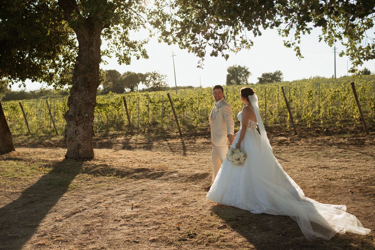 Bride and groom walk hand-in-hand through a sunlit vineyard, bride in elegant white gown with long train.