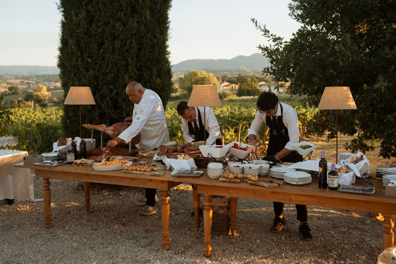 Catering staff serving food at an elegant outdoor buffet table surrounded by cypress trees and scenic countryside views.