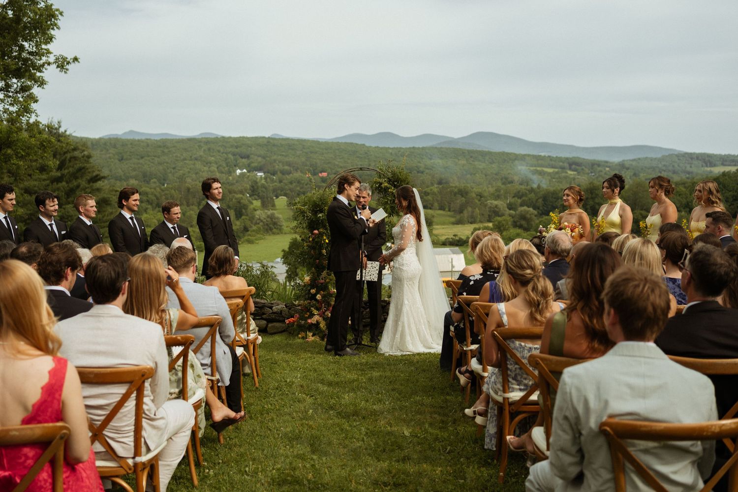Outdoor wedding ceremony with bride and groom at the altar, guests seated on wooden chairs, overlooking a scenic mountain valley.