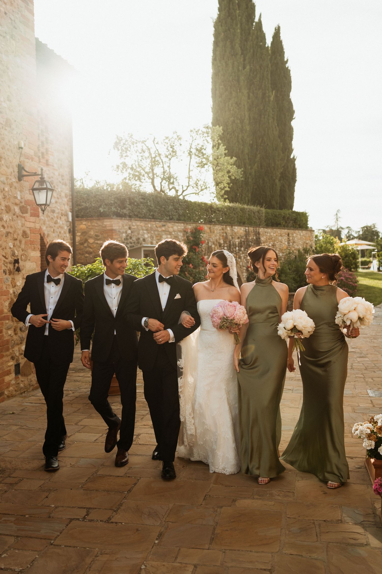 Wedding party walks outdoors at Tuscan venue, bride in white gown with groomsmen in tuxedos and bridesmaids in olive dresses.