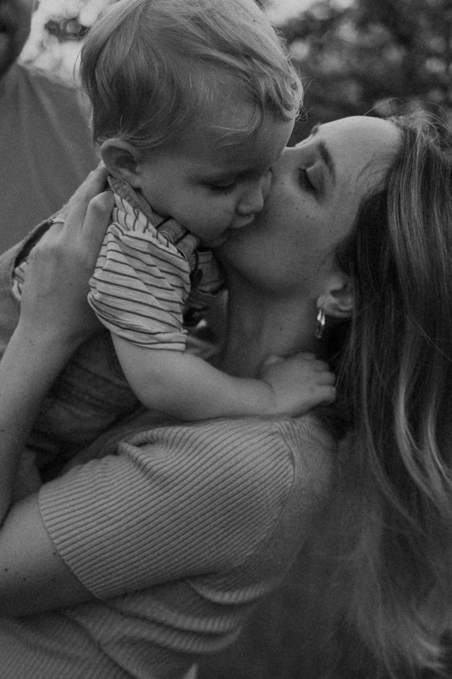 Black and white photo of a mother lovingly kissing her toddler while holding him in her arms outdoors.