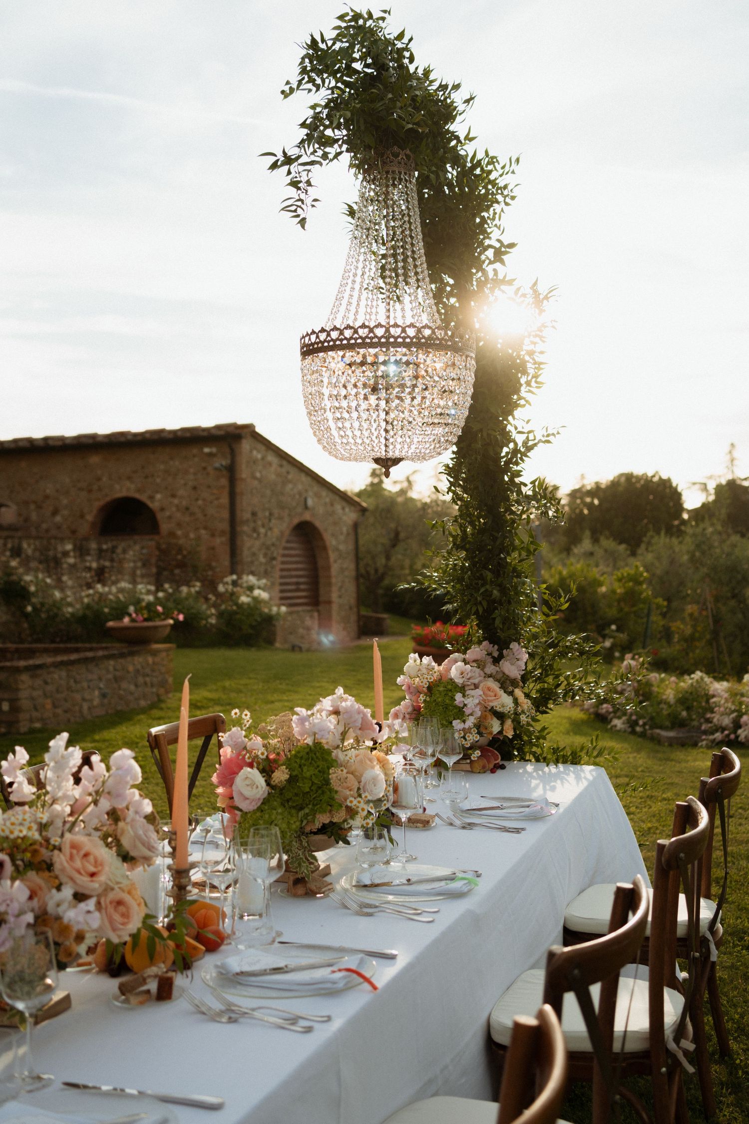 Outdoor Tuscan wedding reception table with crystal chandelier, candles, and floral centerpieces at sunset.