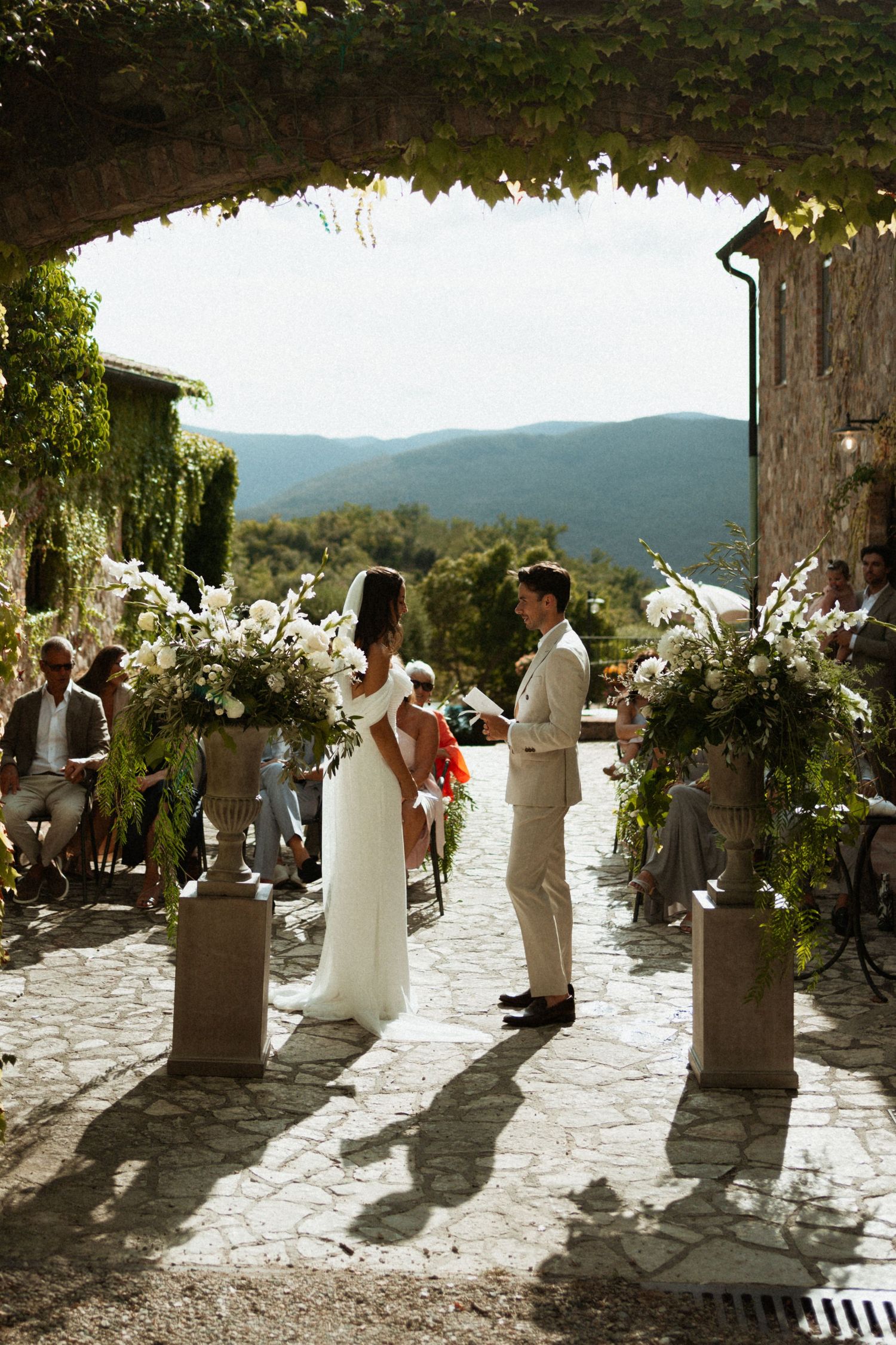 Bride and groom exchanging vows under stone arch with white floral arrangements and mountain views.