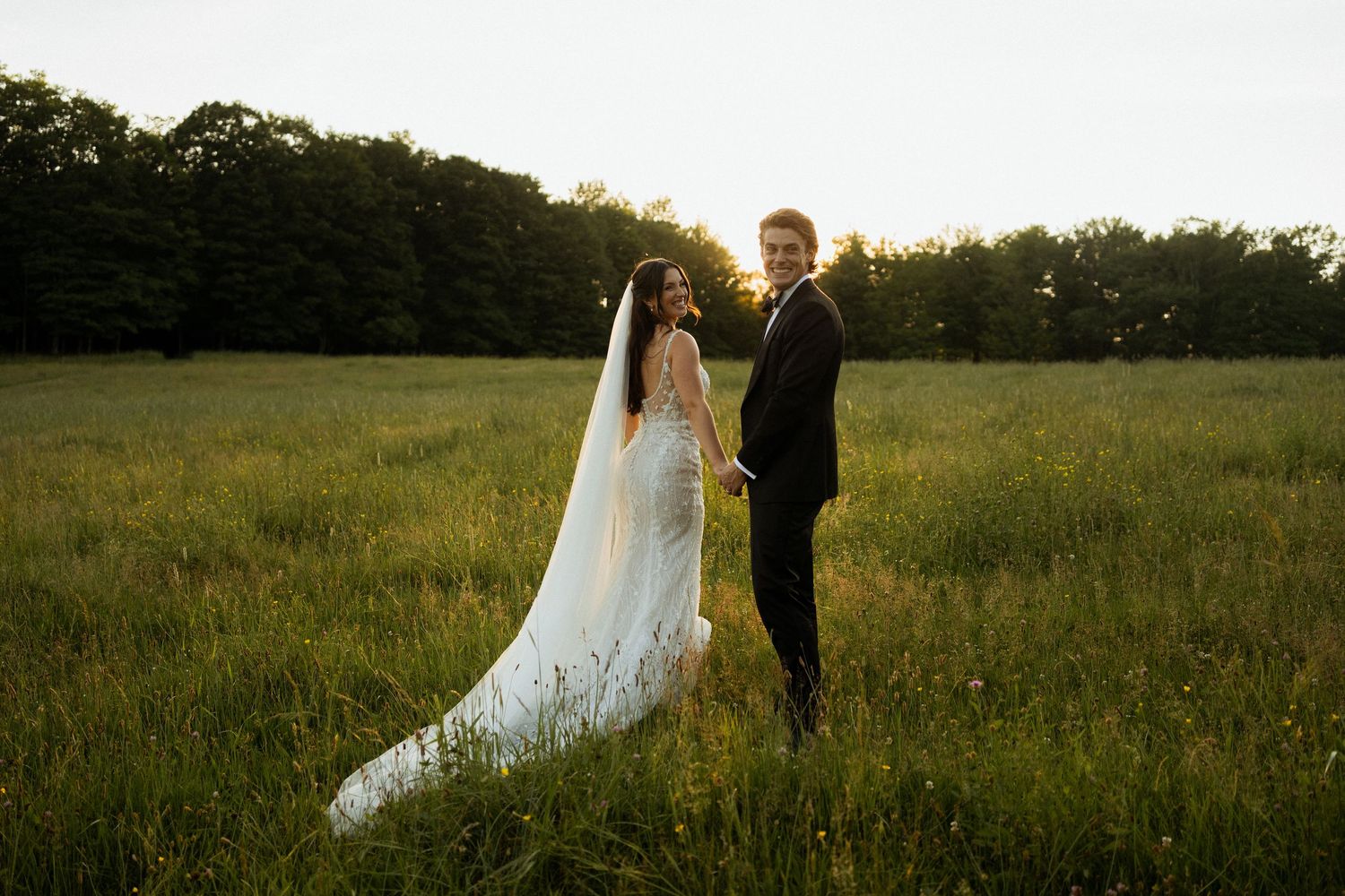 Bride and groom hold hands in golden sunset light across lush green meadow with long veil trailing.