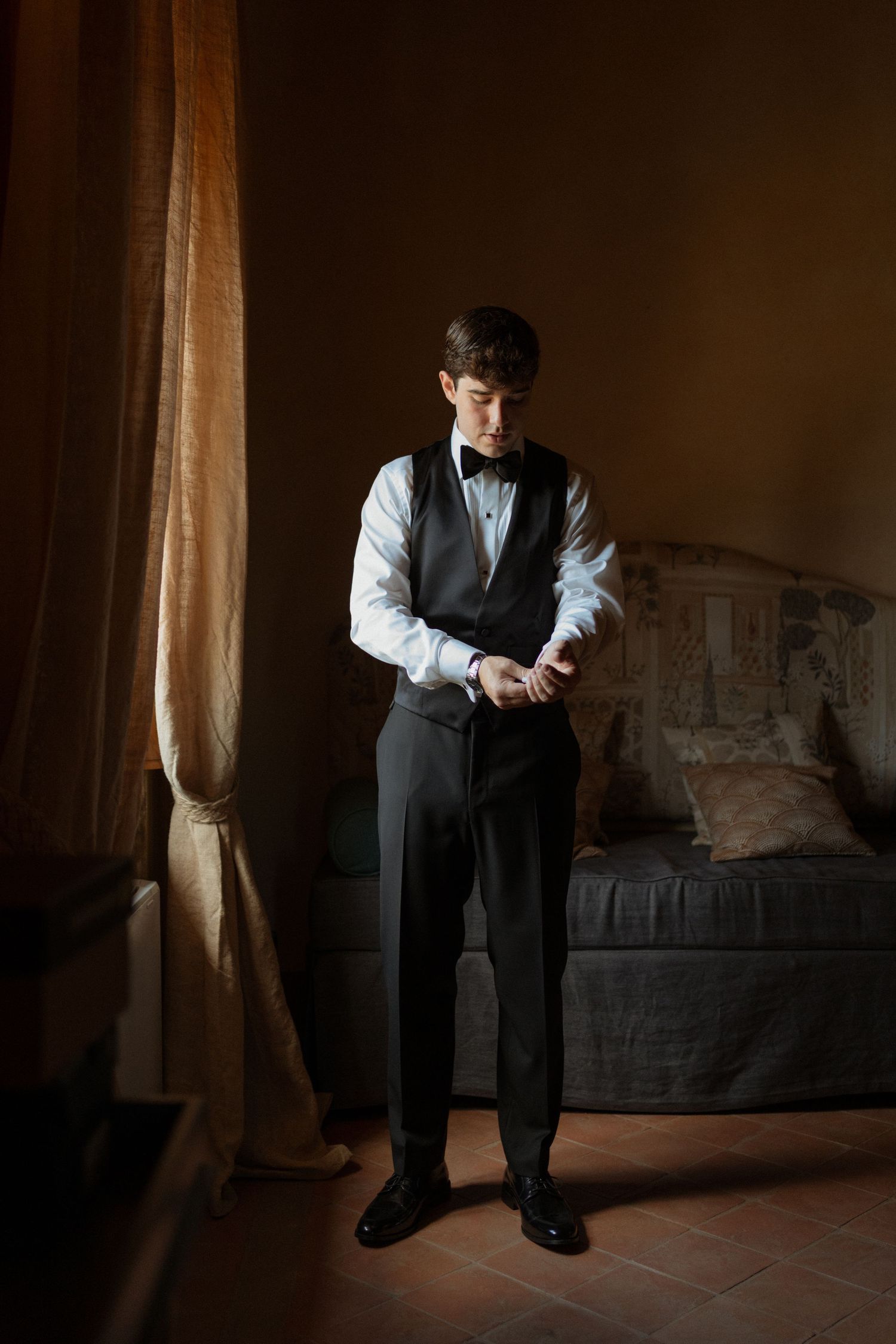 Groom in black tuxedo vest and bow tie adjusts cufflinks in moody, warmly lit hotel room.
