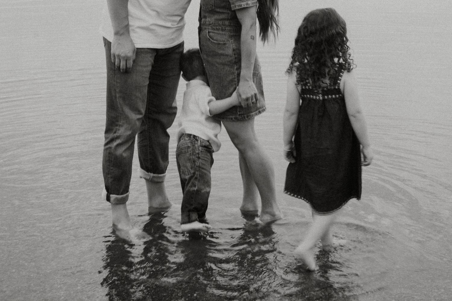 Black and white photo of a family standing in shallow water, viewed from behind, with two young children.