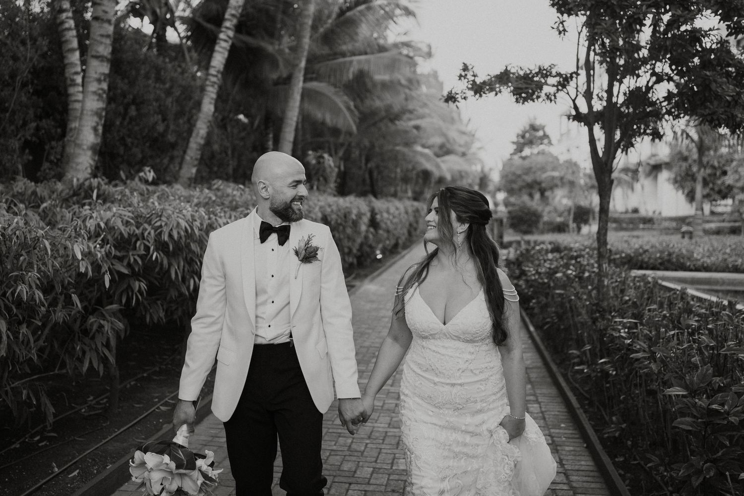 Black and white photo of a bride and groom holding hands, walking along a garden path surrounded by tropical trees.