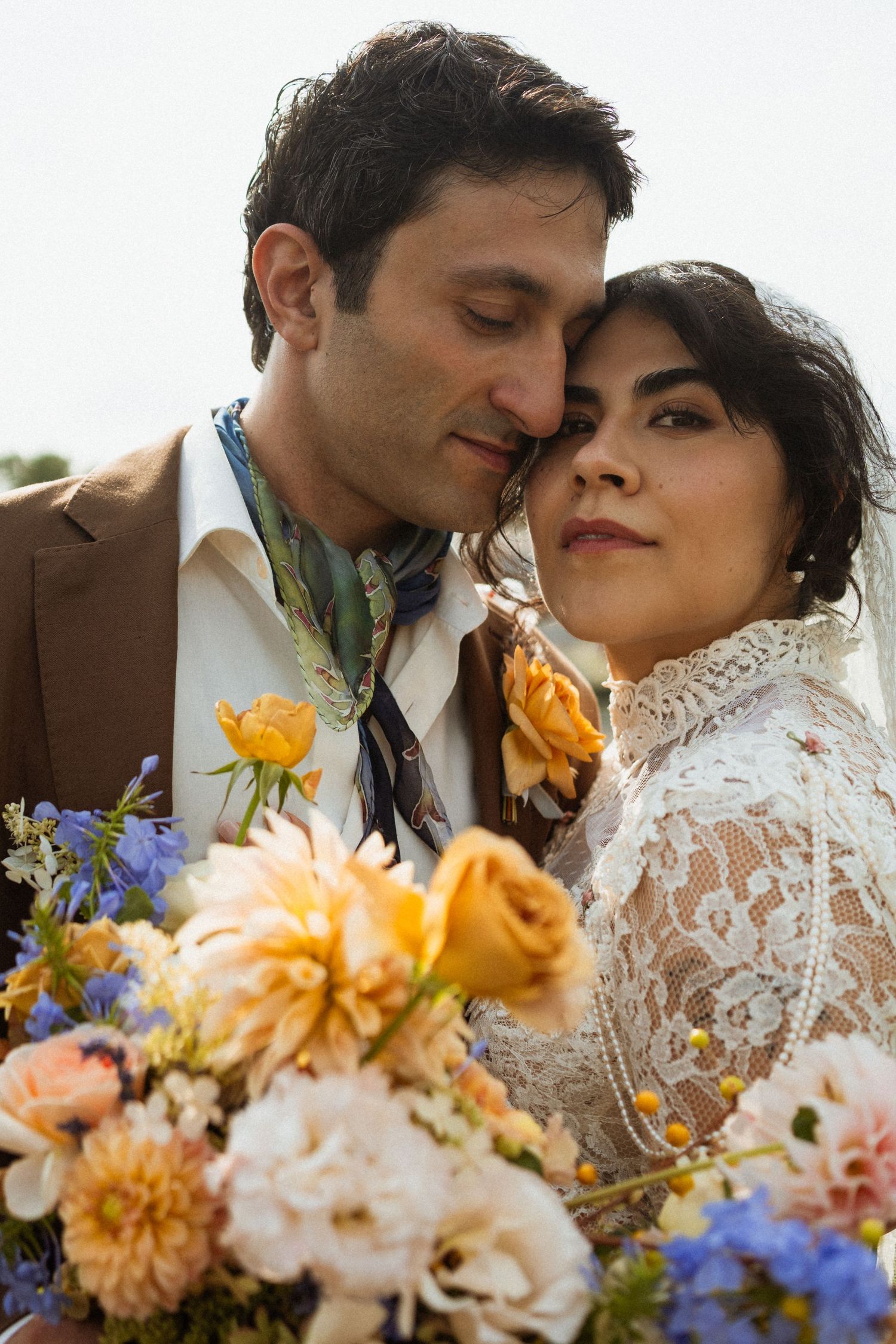 Romantic wedding couple in lace dress and brown suit with colorful floral bouquet of yellow roses and blue flowers.