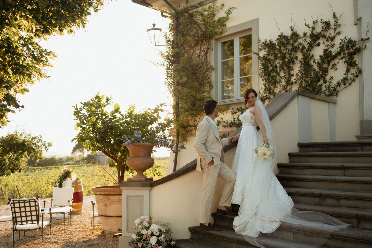 Bride and groom holding hands on stone steps outside a rustic villa surrounded by lush greenery on their wedding day.