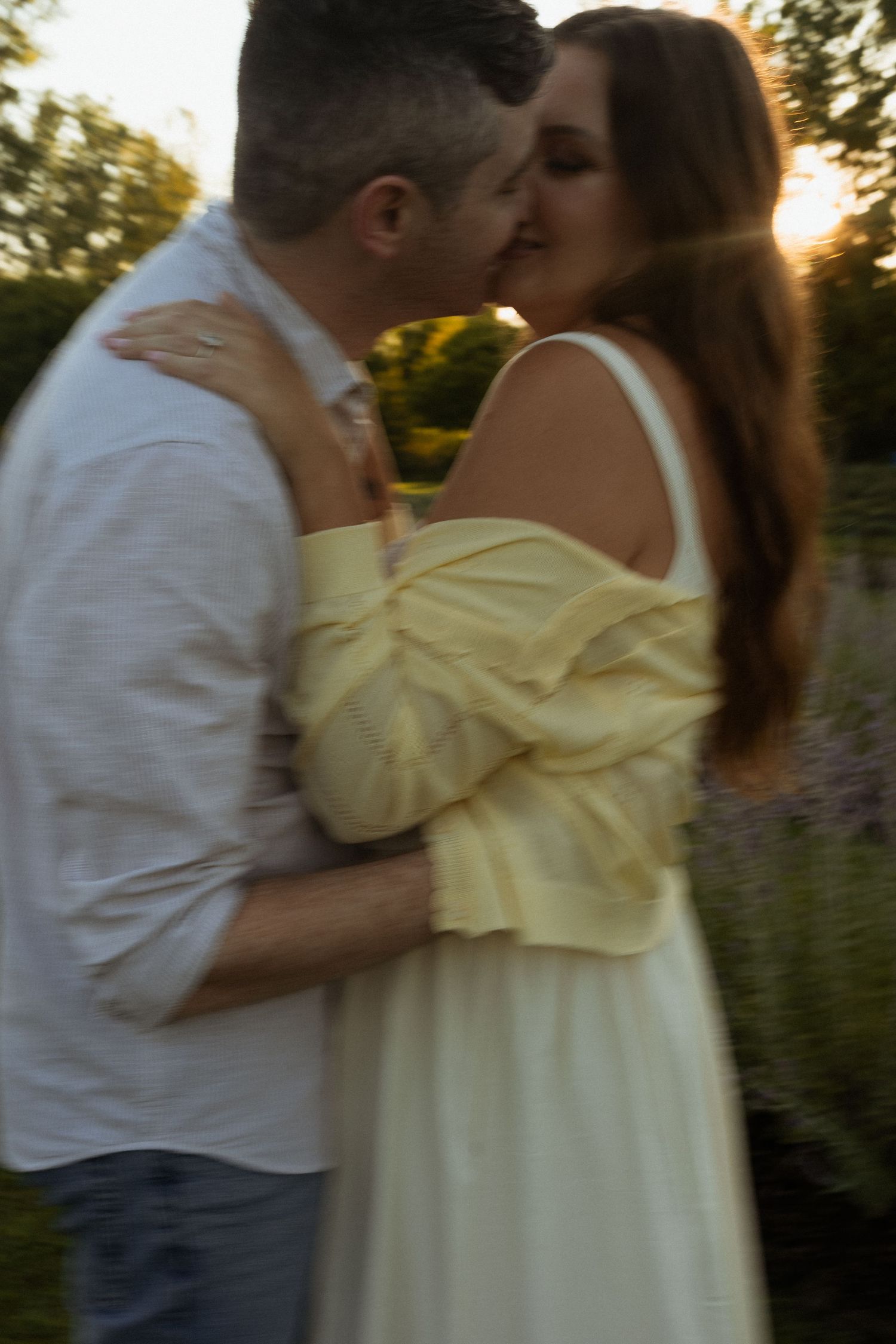 A couple shares a romantic kiss at sunset, woman in yellow off-shoulder dress embracing man in white shirt outdoors.