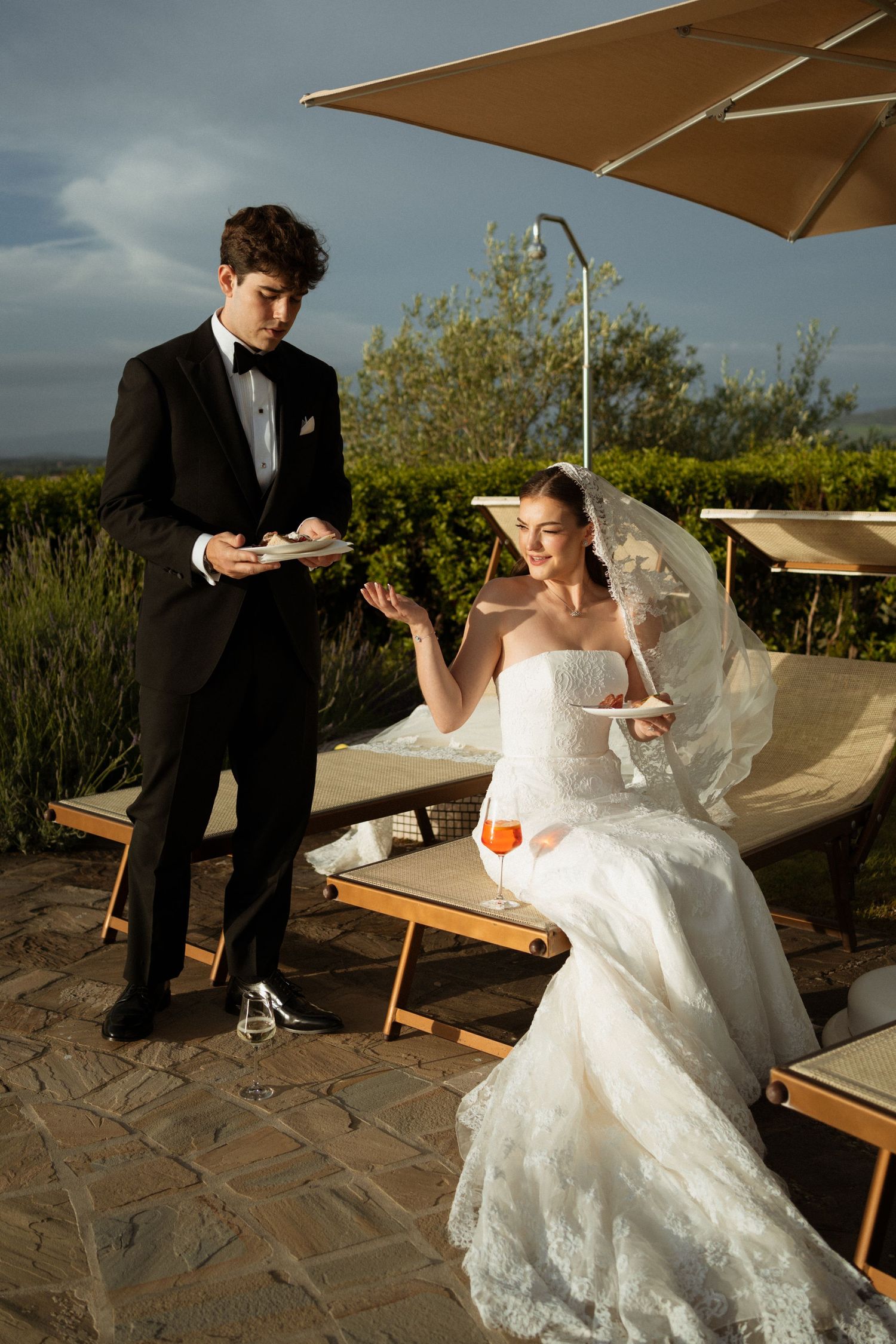 Bride in white lace gown with veil sits outdoors holding drink while groom in black tuxedo stands beside her.