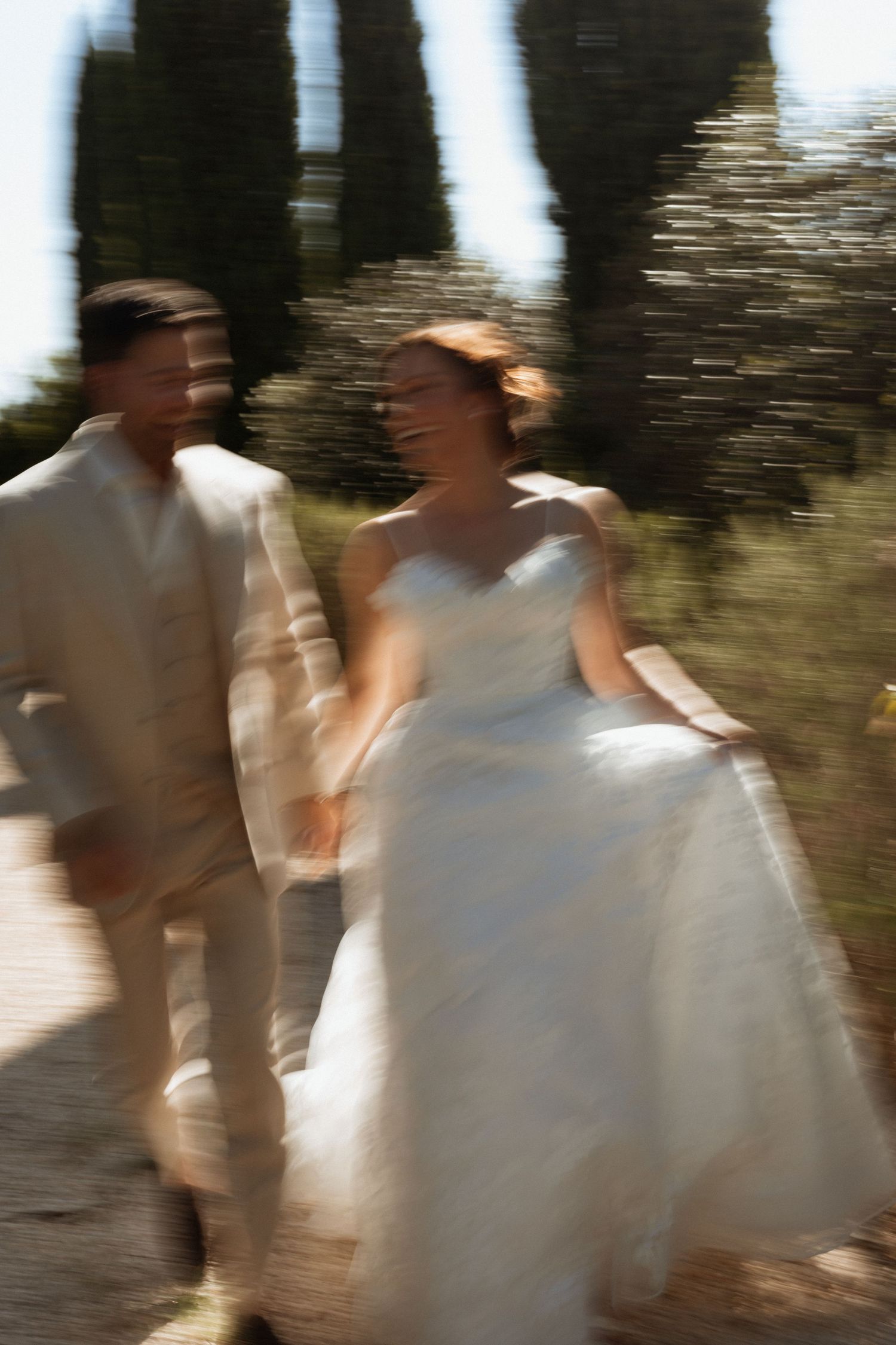 Blurred motion shot of smiling bride in white gown and groom in beige suit walking hand in hand outdoors.