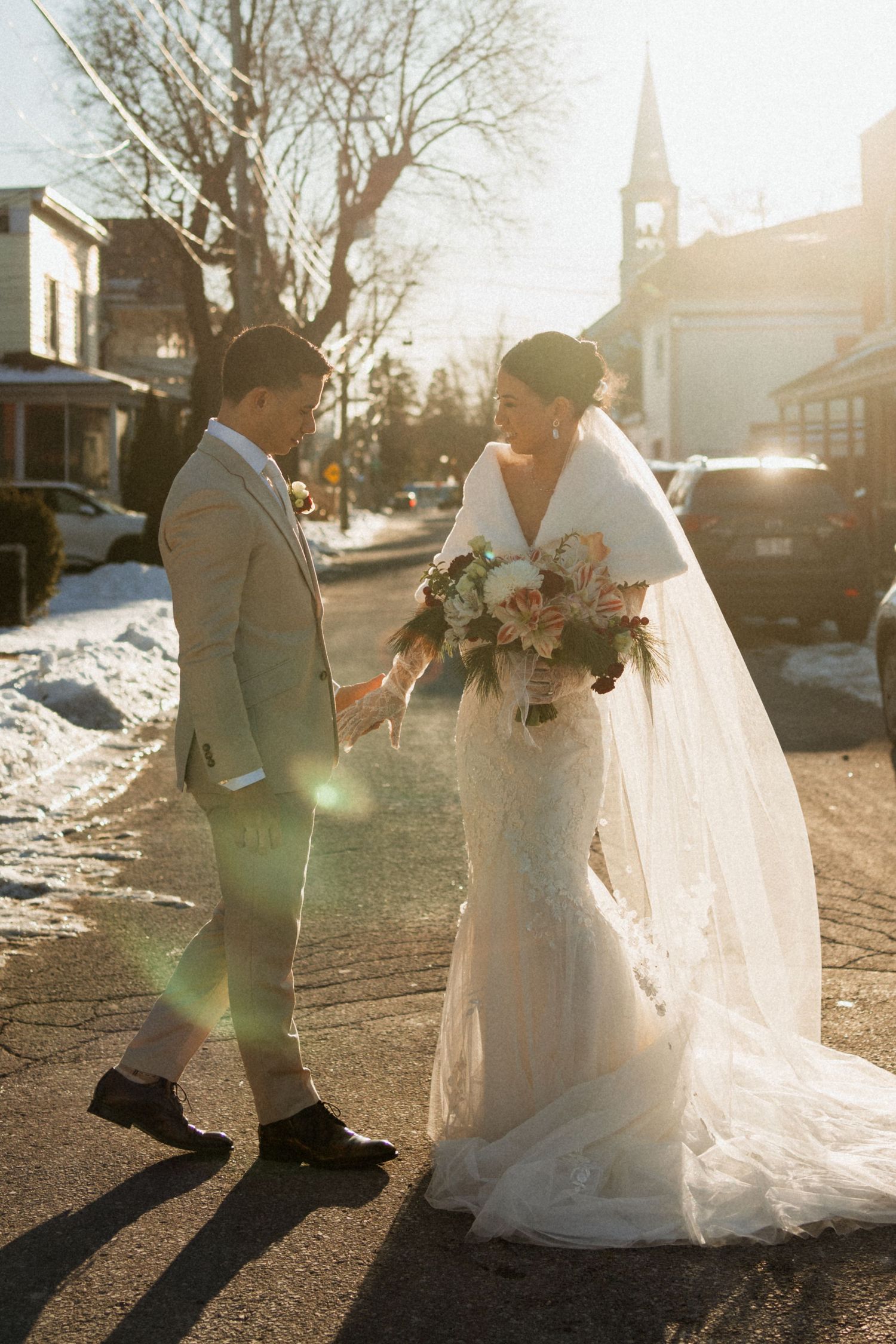 Bride and groom holding hands on snowy street in golden winter sunlight outside church wedding venue.