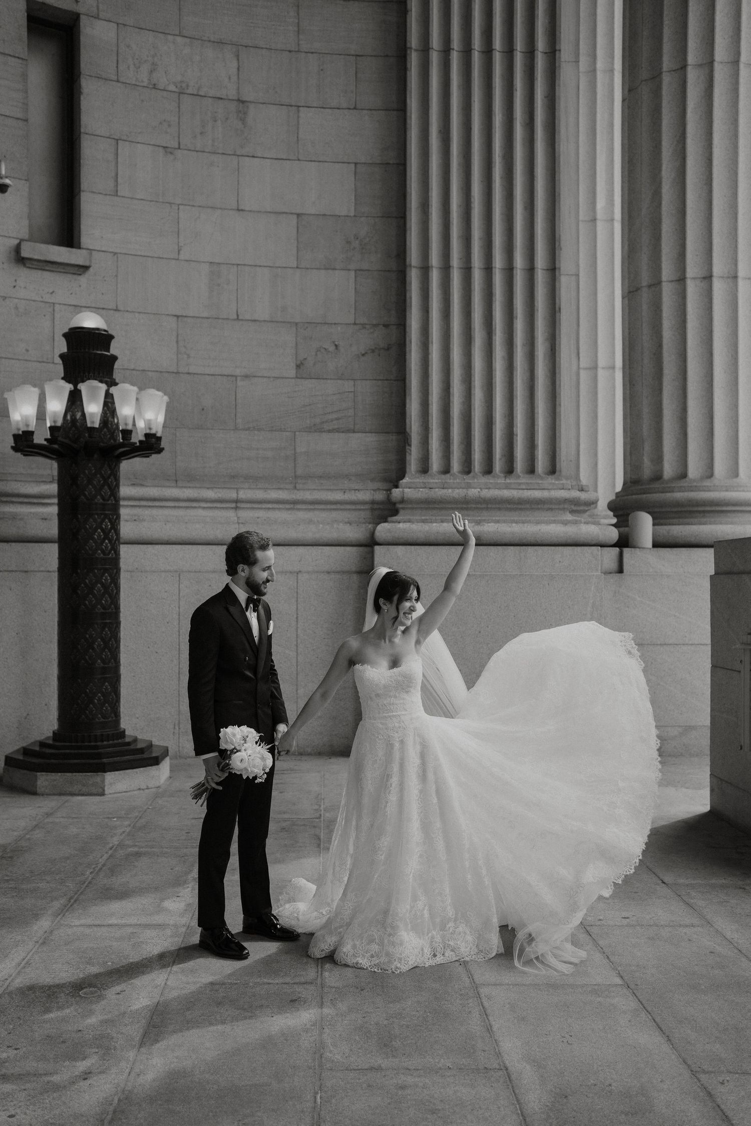 Black and white photo of bride twirling gown beside groom near grand stone columns.
