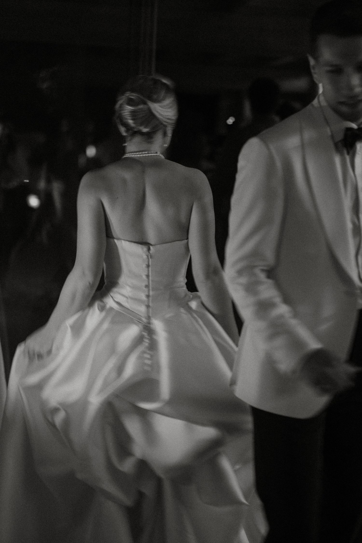 Elegant bride in strapless backless white satin gown with button detail walks beside groom in white tuxedo.