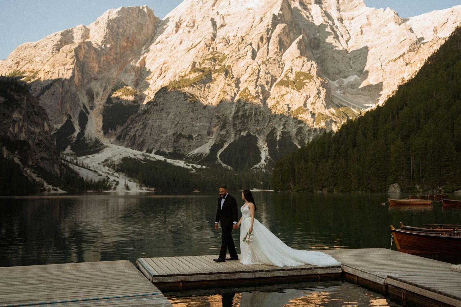 Bride and groom standing on wooden dock with stunning alpine lake and mountain backdrop at sunset.