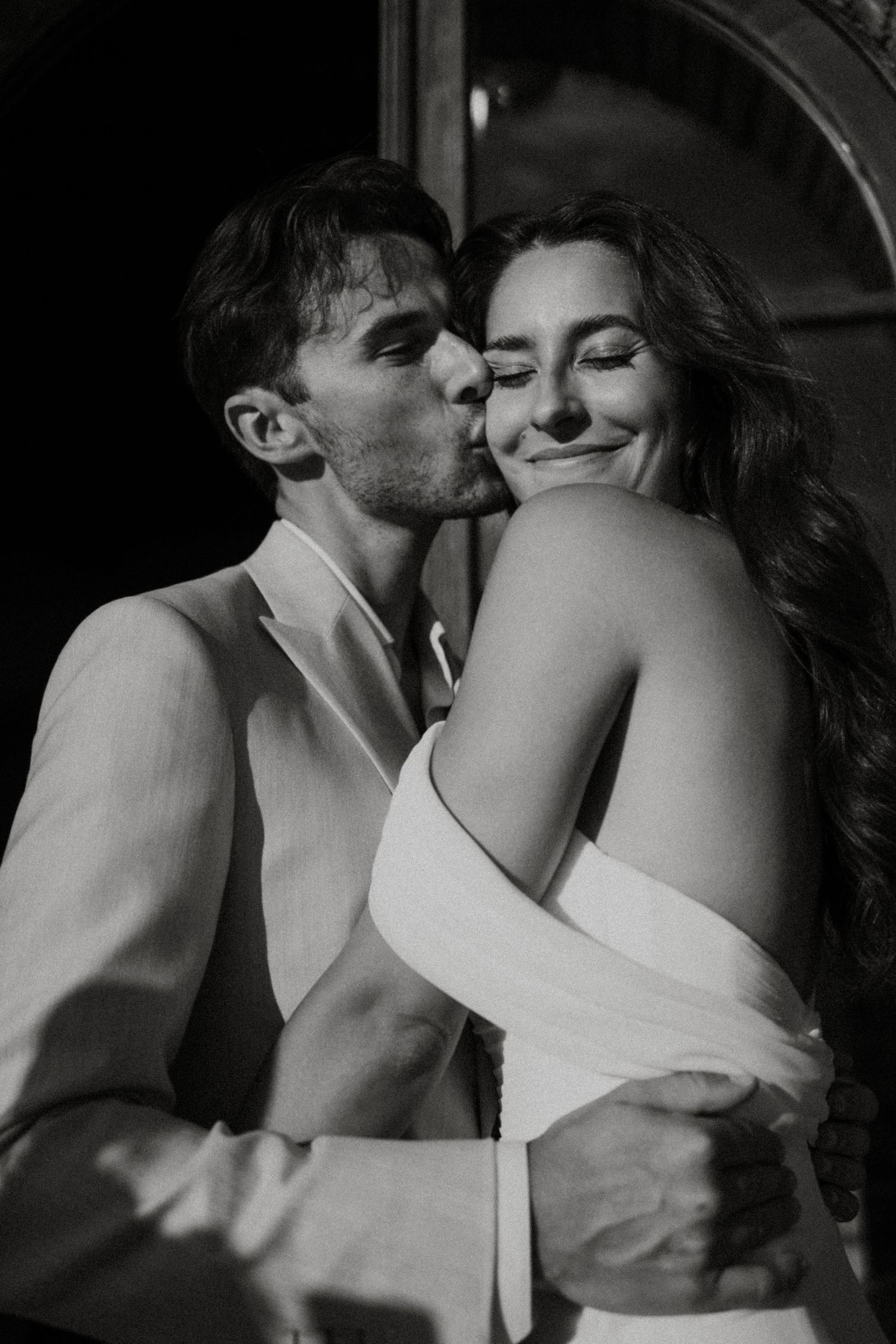 Romantic black and white photo of groom kissing bride on cheek while embracing her from behind.