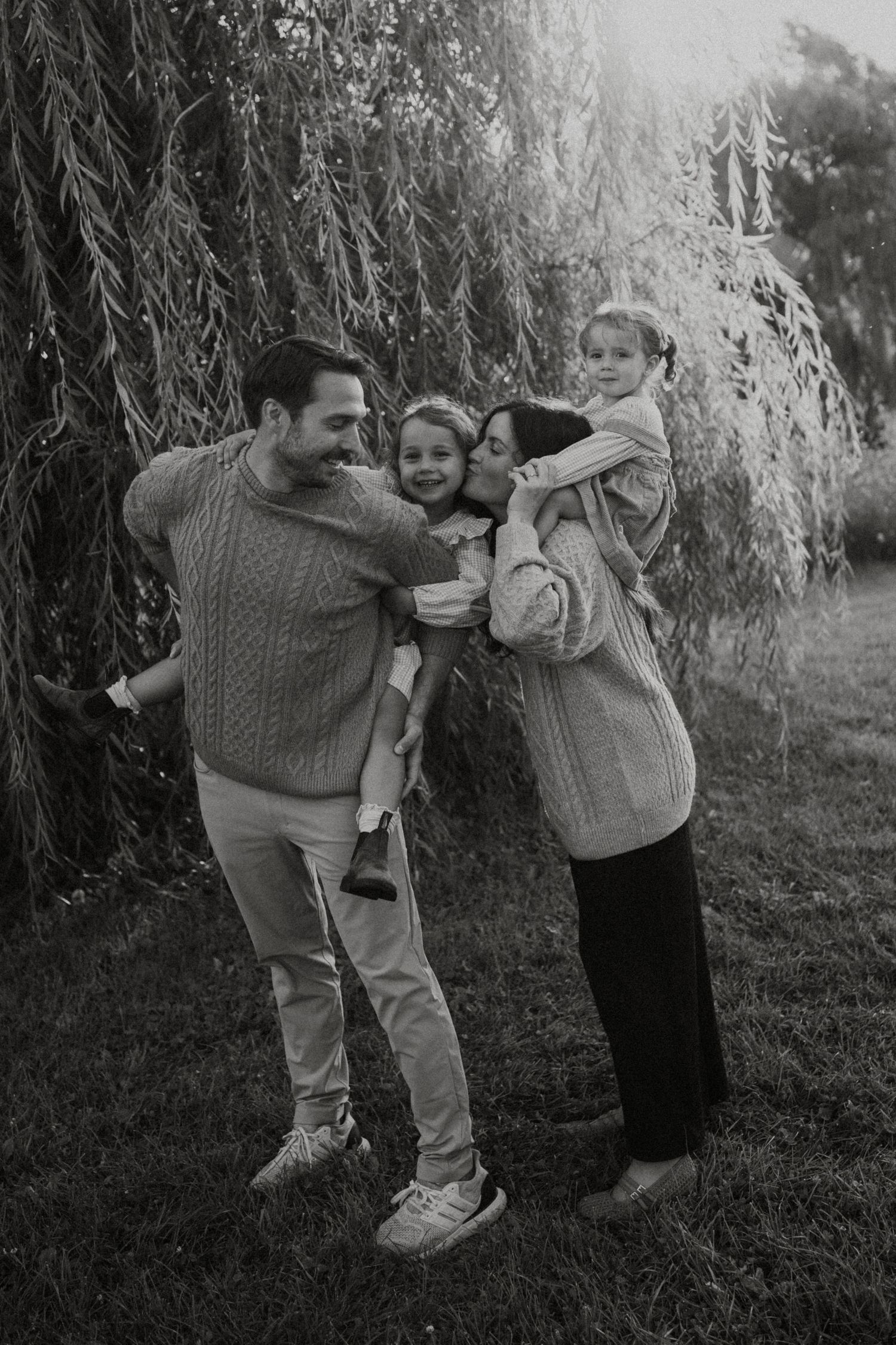 Black and white family portrait of parents holding two young children outdoors near a weeping willow tree.