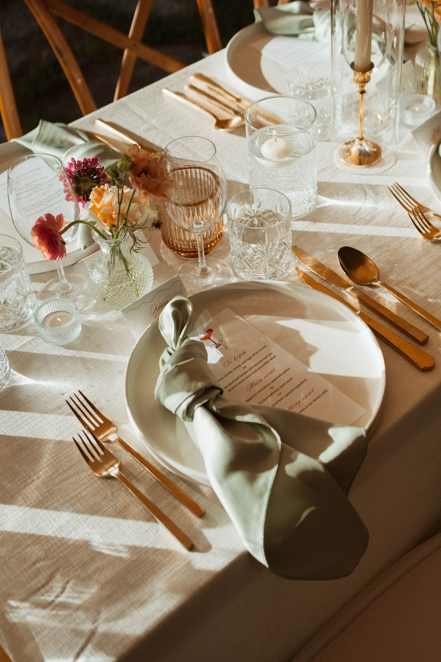 Elegant wedding table setting with gold cutlery, sage green napkin, crystal glasses, and colorful floral centerpiece in warm sunlight.