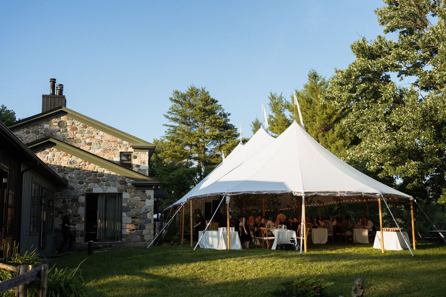 Elegant white sailcloth tent set up on green lawn beside stone building for outdoor wedding reception.