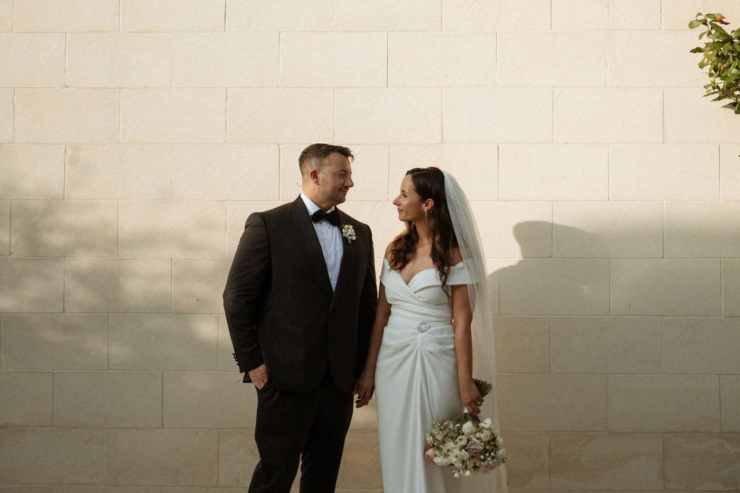 Bride and groom sharing a tender moment standing against a sunlit white brick wall.
