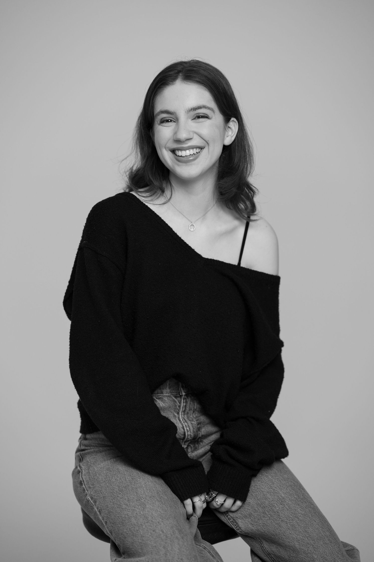Smiling young woman wearing black off-shoulder sweater poses in bright black and white studio portrait.
