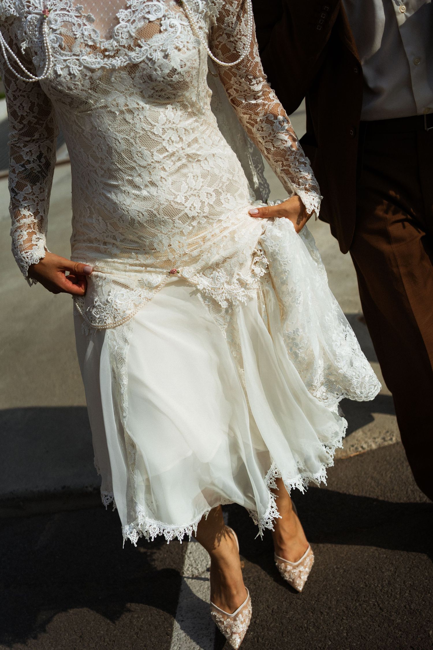 Bride in elegant white lace wedding dress with long sleeves and lace heels, walking with groom on sunny day.