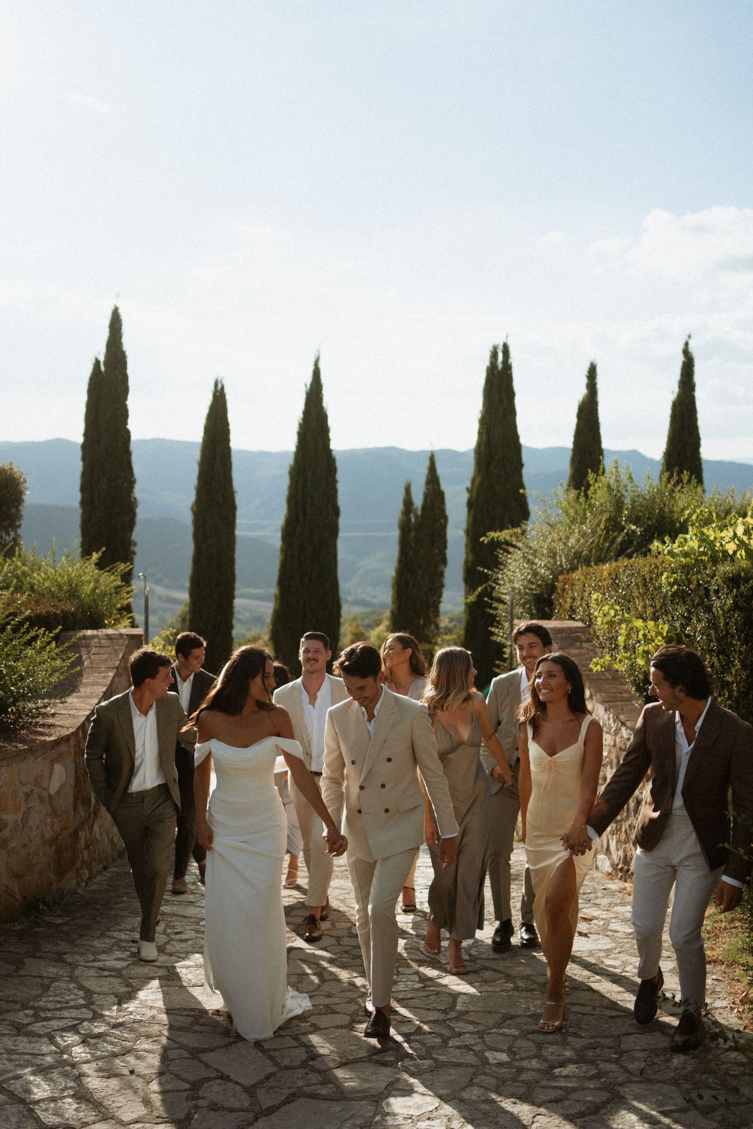 Wedding party walking on stone path with cypress trees and Tuscan hills backdrop on sunny day.