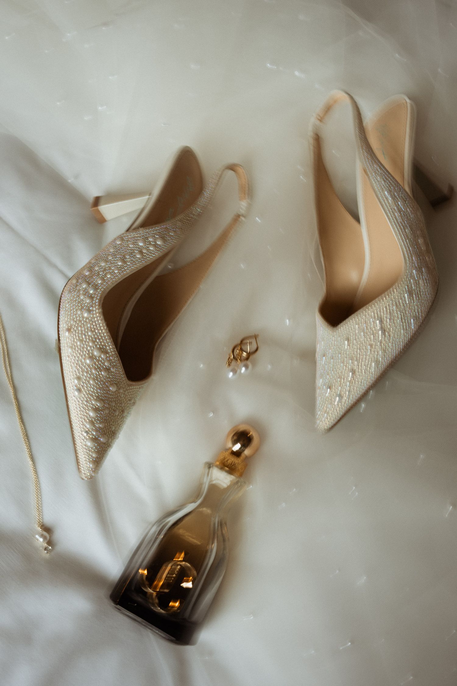 Sparkly ivory wedding heels with pearl earrings and gold perfume bottle on white fabric.