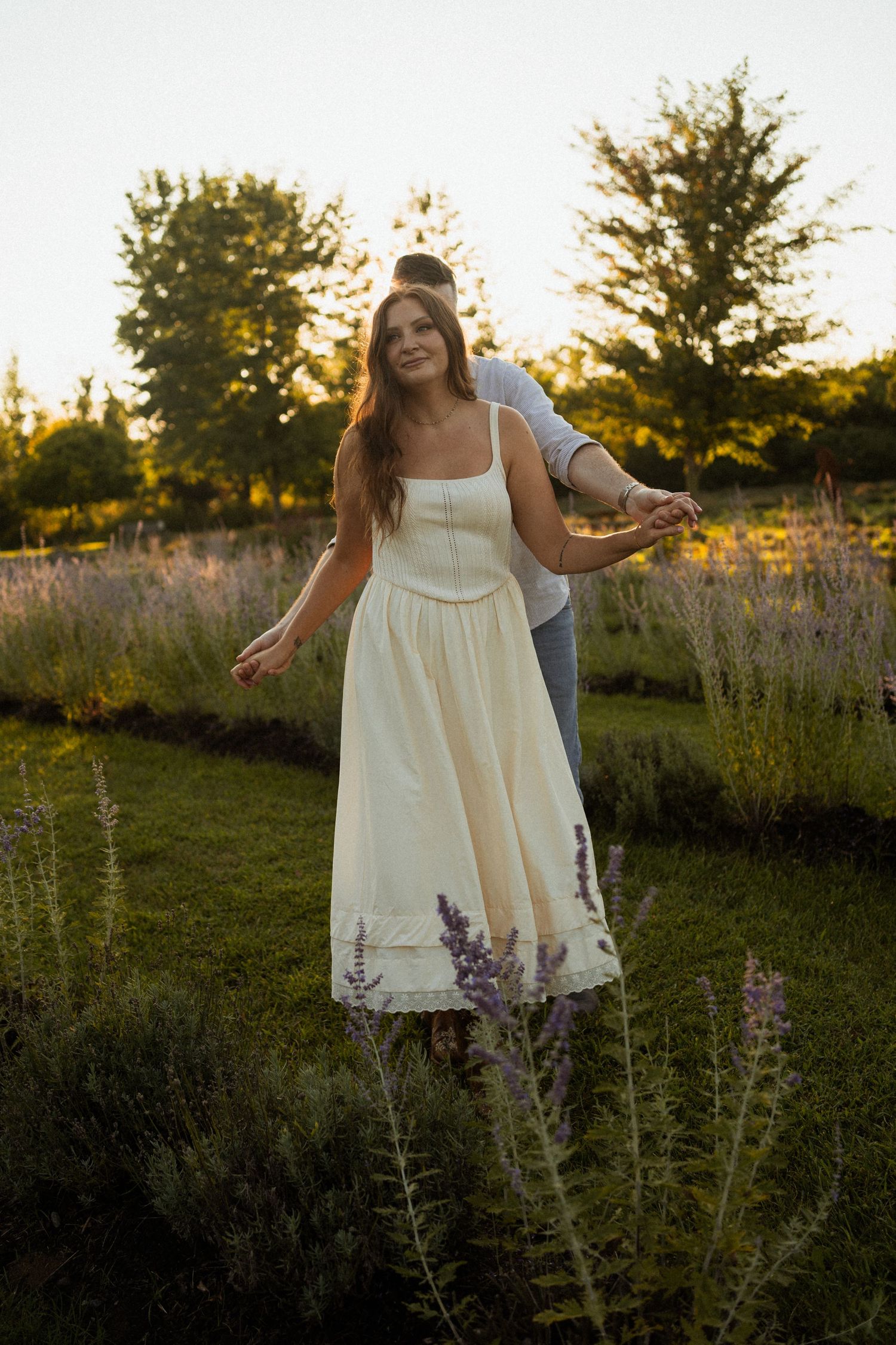 Couple in lavender field at golden hour, woman in cream dress holding hands with man behind her.