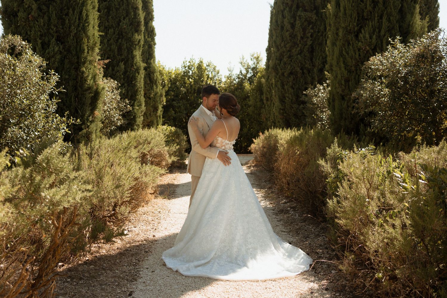 Bride and groom kissing on a garden path lined with cypress trees and olive shrubs on their wedding day.
