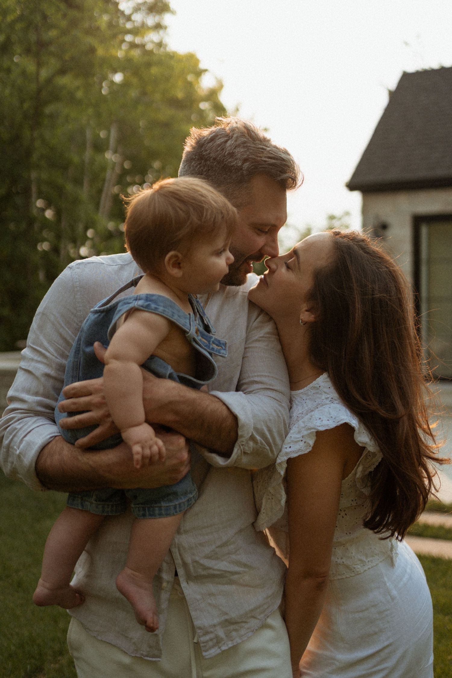 A happy young family with a baby outdoors, parents sharing a kiss while holding their infant child near a house.
