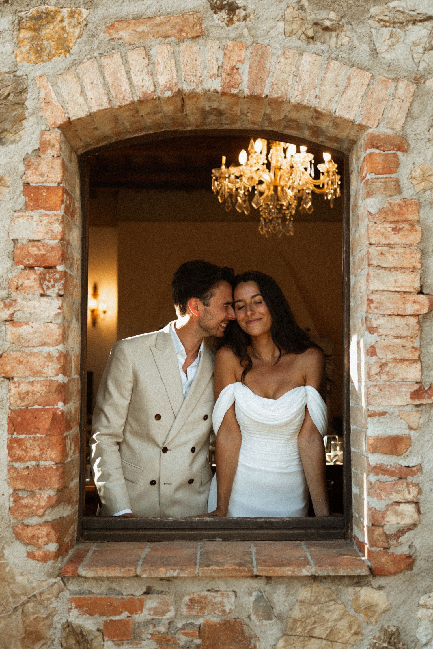 Bride and groom sharing romantic moment framed in arched stone window with chandelier backdrop.
