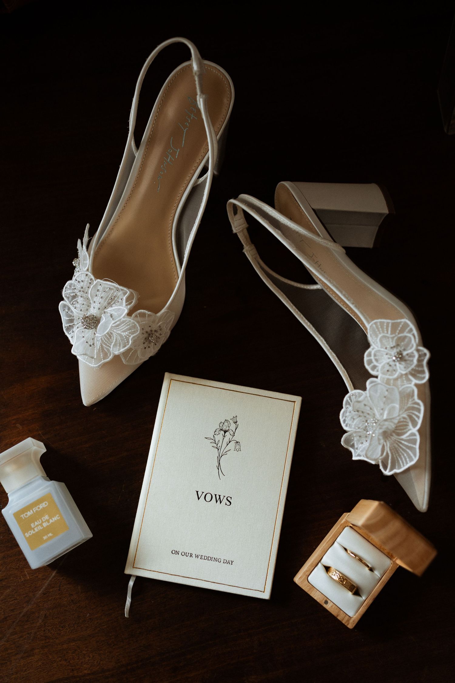 Wedding flat lay with lace heels, vow book, rings, and perfume on dark wood.