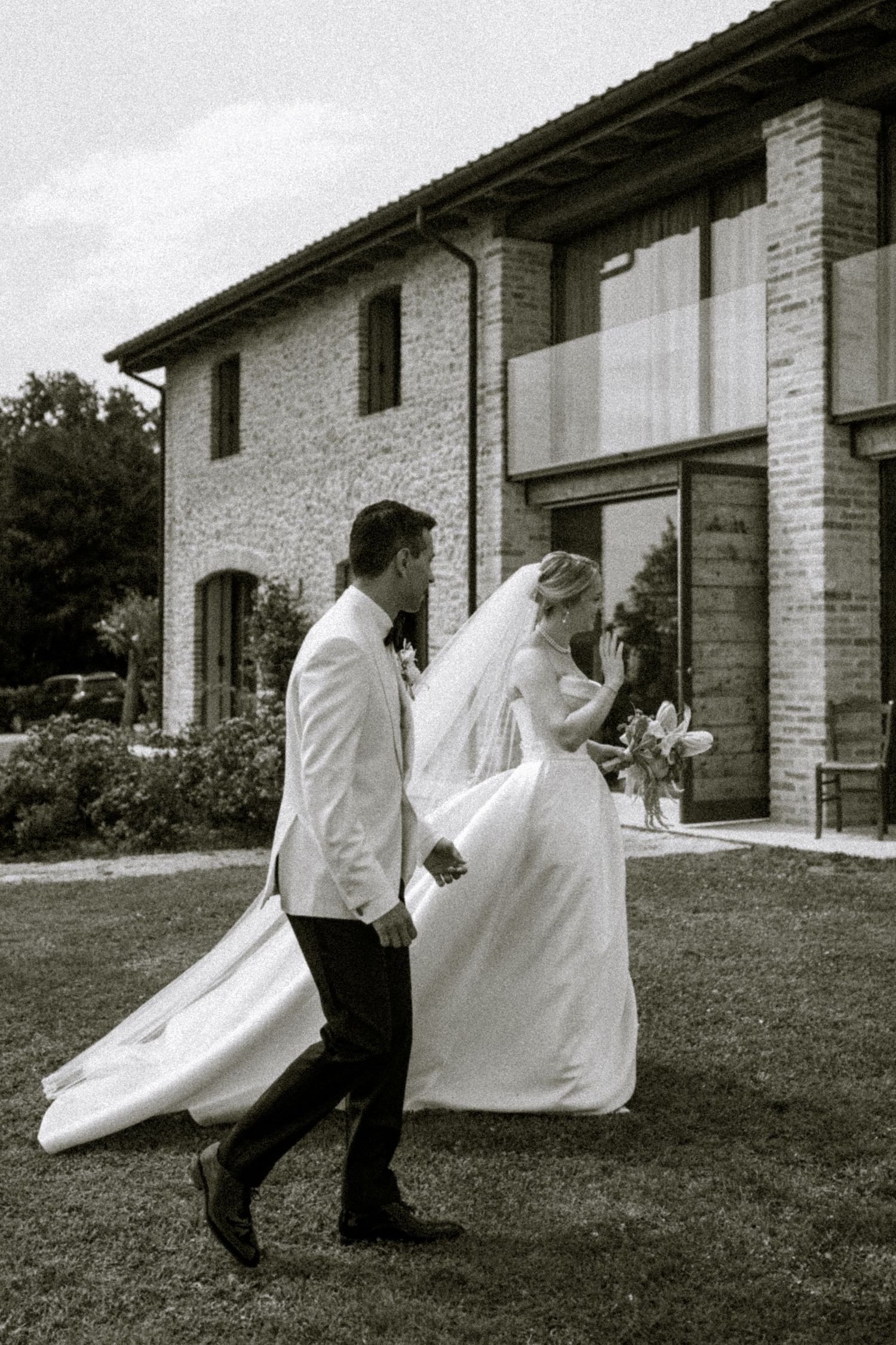 Black and white photo of bride and groom walking across lawn outside rustic stone villa on wedding day.