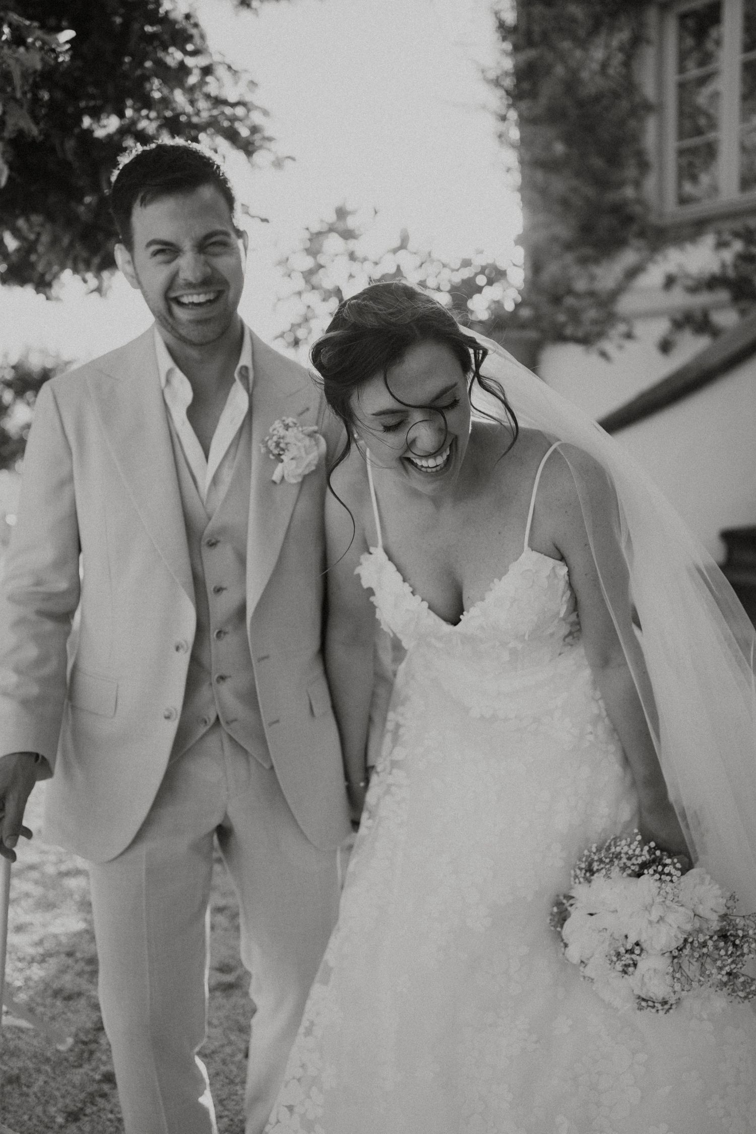 Black and white photo of a laughing bride and groom walking together on their wedding day, bride holding a bouquet.