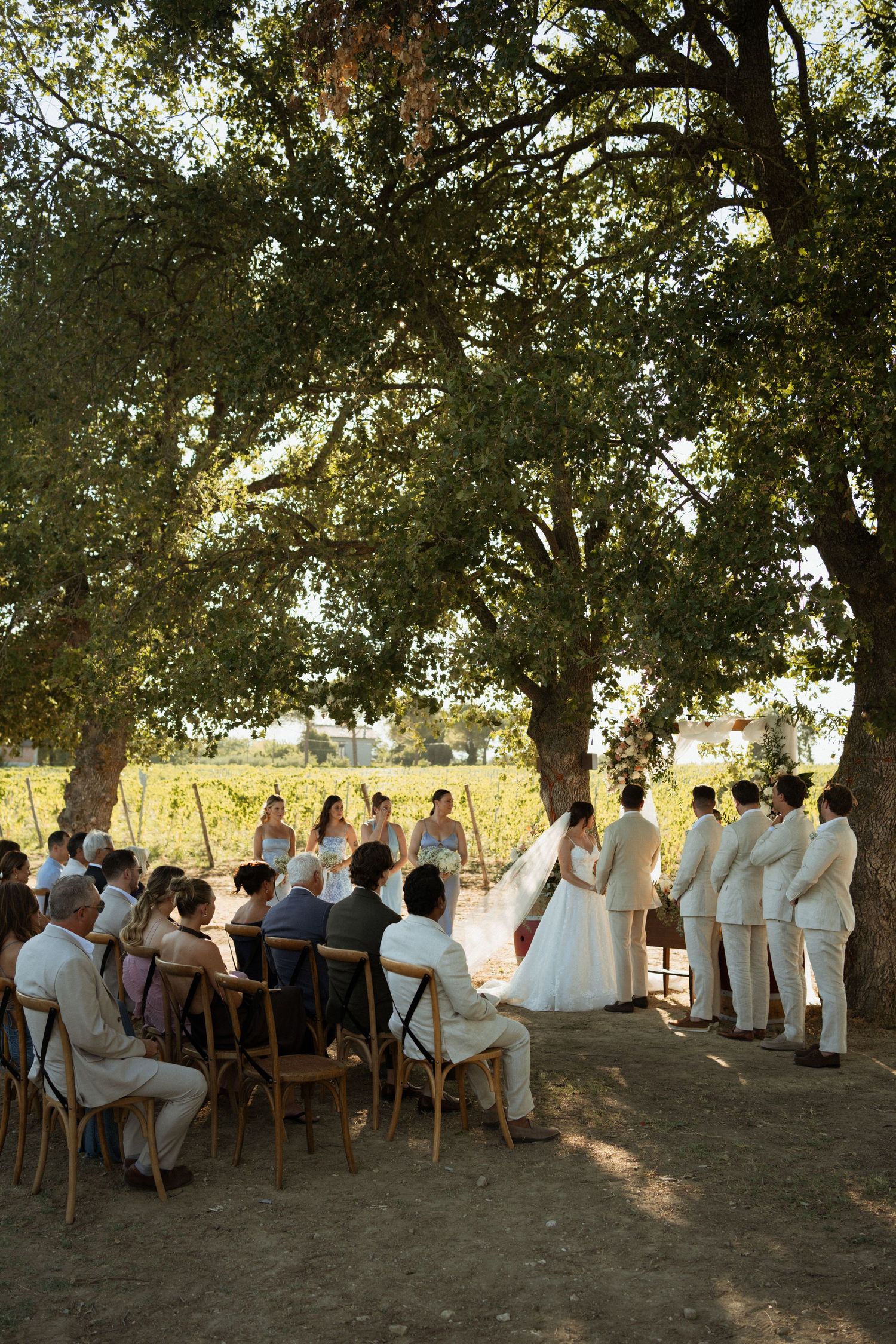 Outdoor wedding ceremony under oak trees at a vineyard, with guests seated and wedding party standing in golden sunlight.
