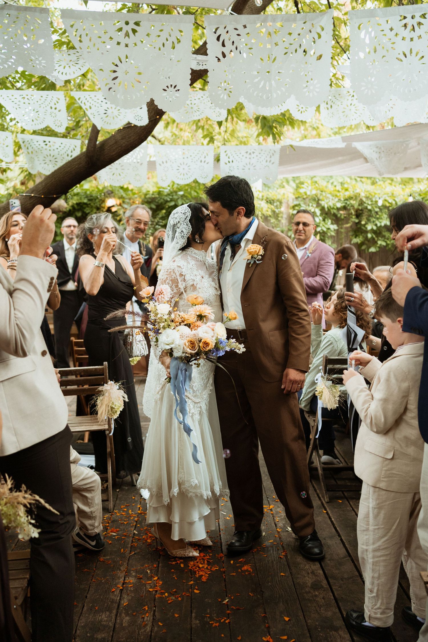 Bride and groom share a kiss at outdoor wedding ceremony with papel picado decorations and guests celebrating around them.
