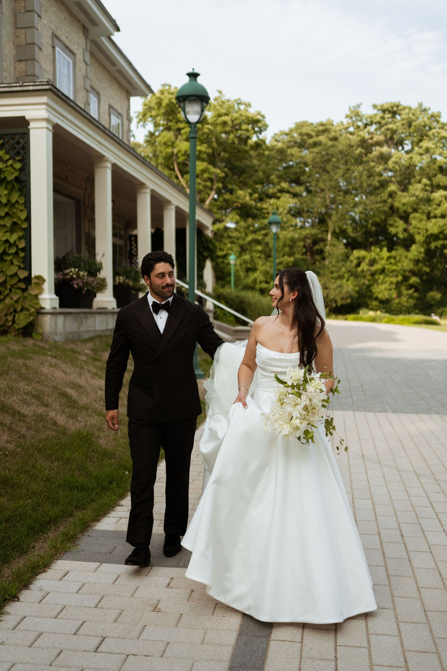 A bride and groom walk hand-in-hand along a brick path outside an elegant mansion on their wedding day.
