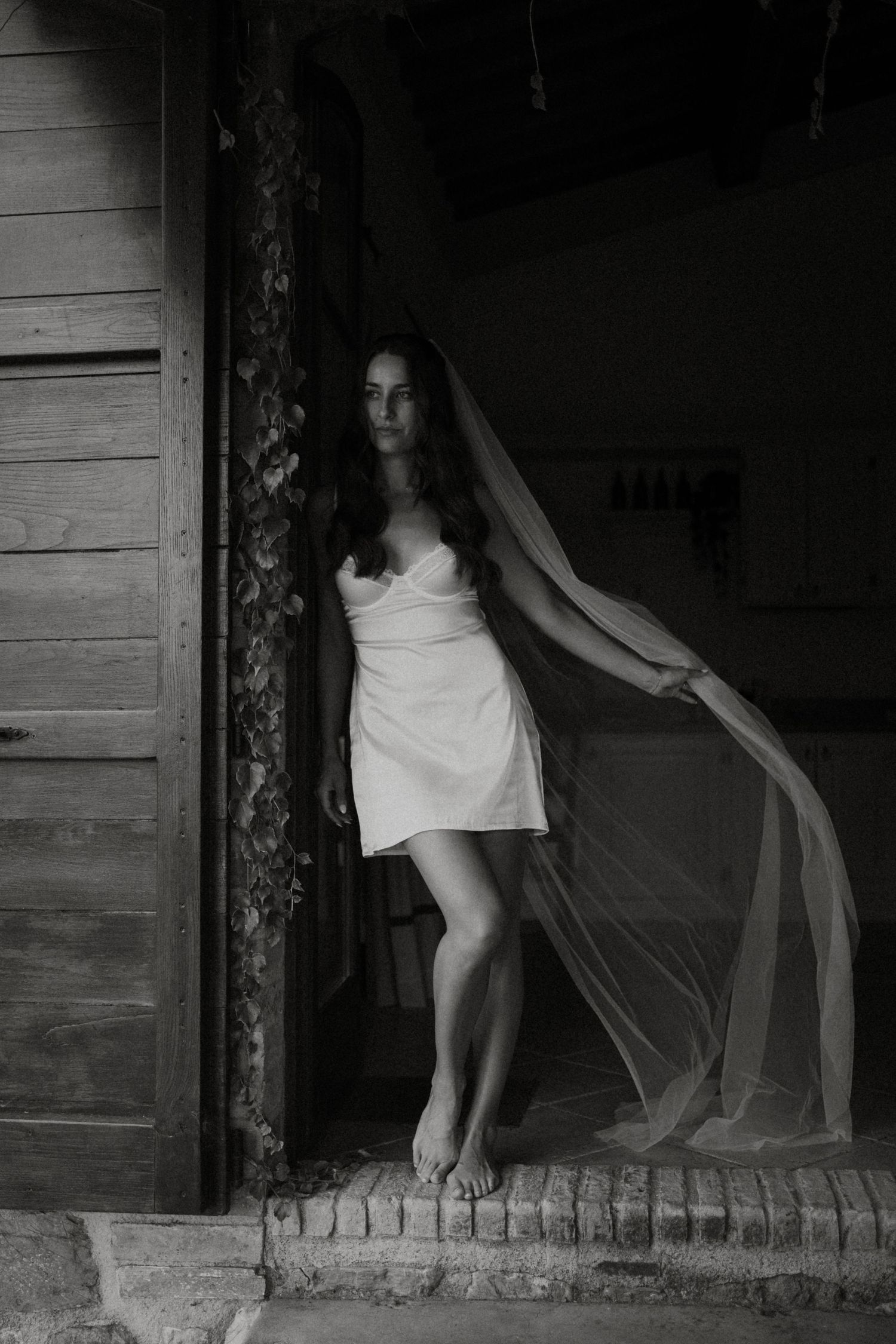Black and white portrait of bride in short white dress holding flowing veil in doorway.