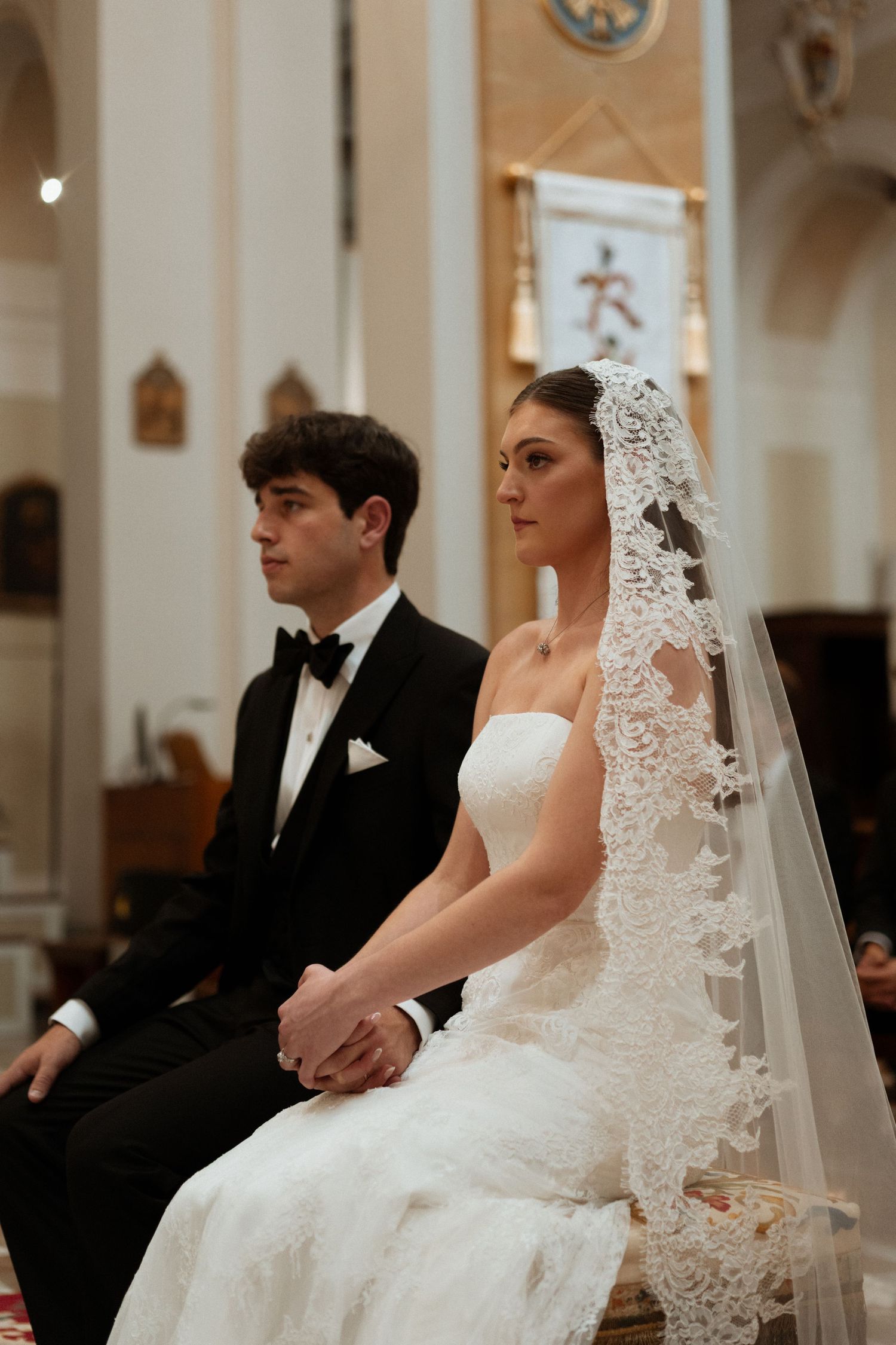 Bride and groom seated during church wedding ceremony, bride wearing lace mantilla veil.